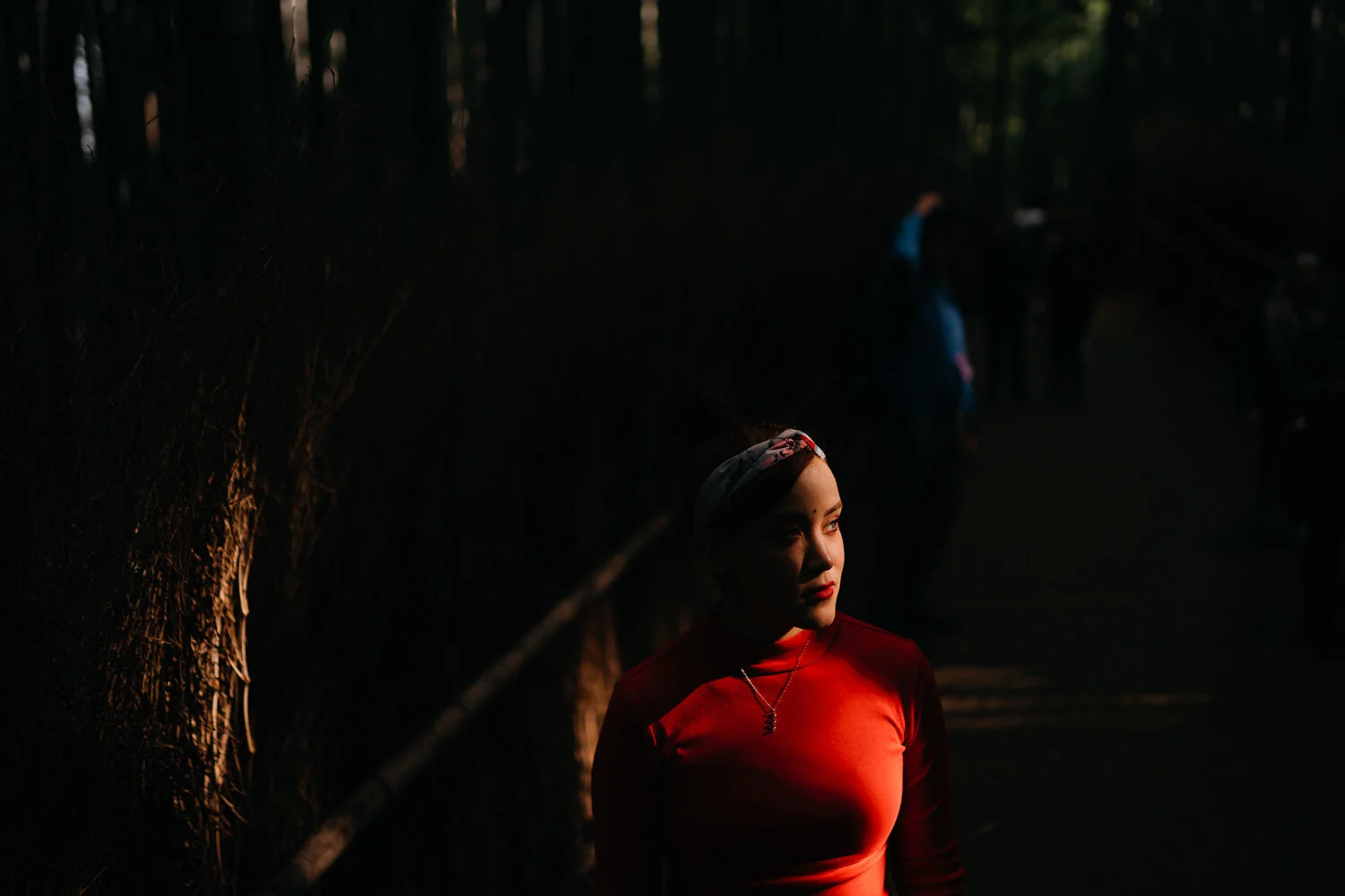 A woman with makeup and a headscarf in a red shirt standing in a dark forest, illuminated by sunlight.