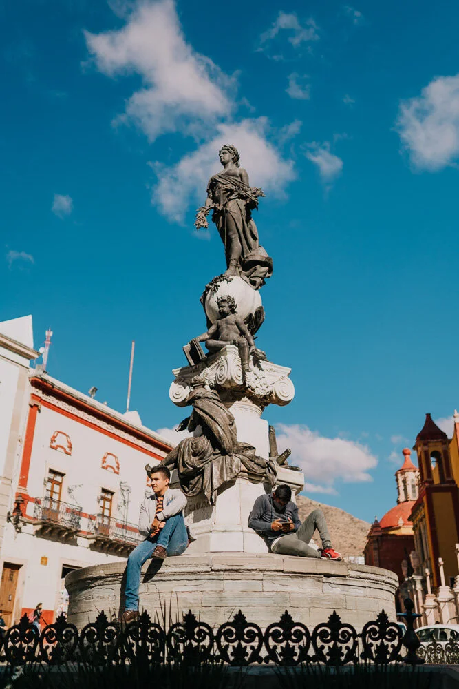 A tall, ornate statue with multiple figures on top, set in a city square with surrounding buildings and a mountain in the distance, under a blue sky with clouds.