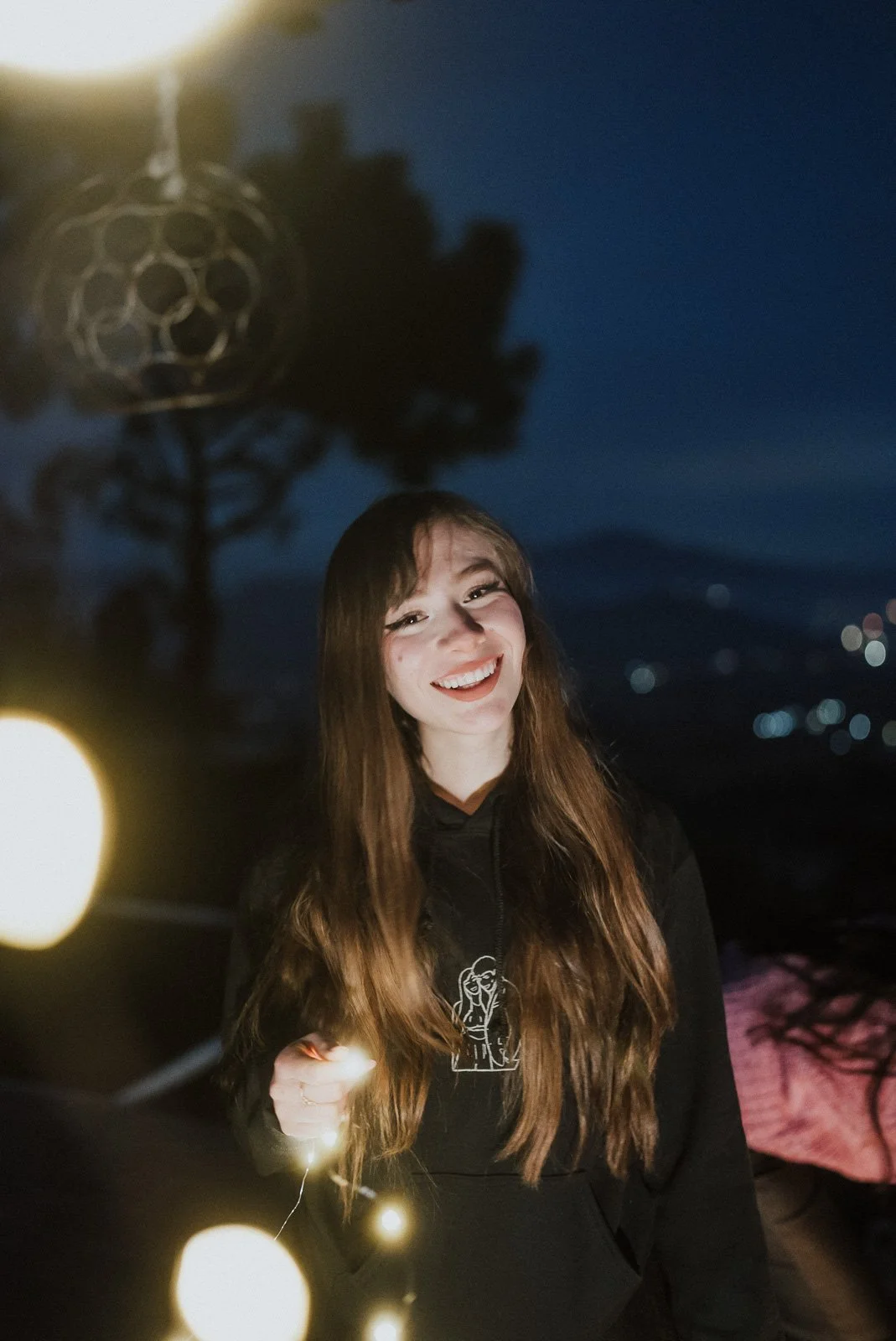 A young woman with long brown hair, smiling and holding a string of fairy lights in a dark outdoor setting at night.