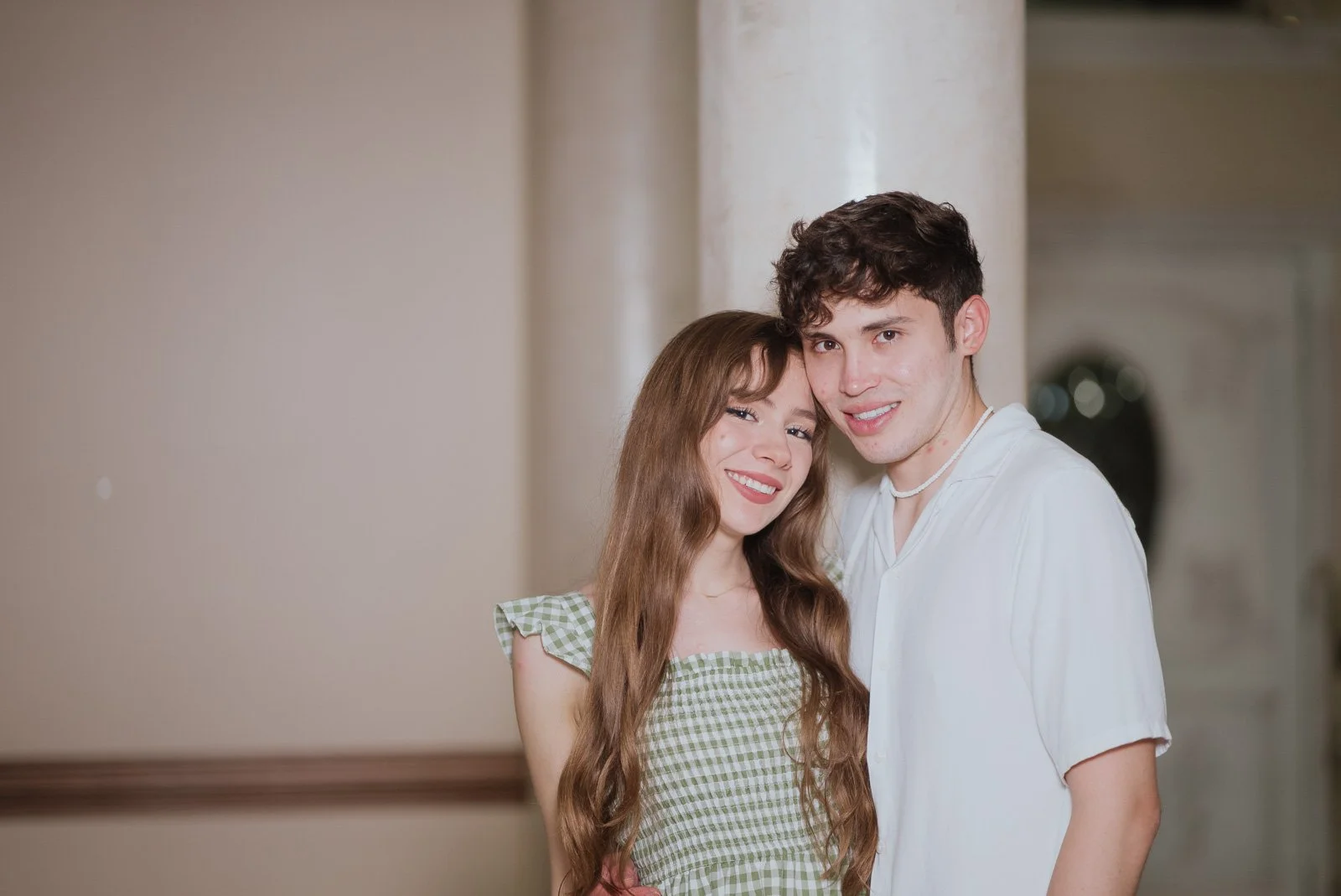 A smiling young woman with long wavy hair and a young man with short curly hair standing close together indoors, with neutral-colored walls and a blurred background.