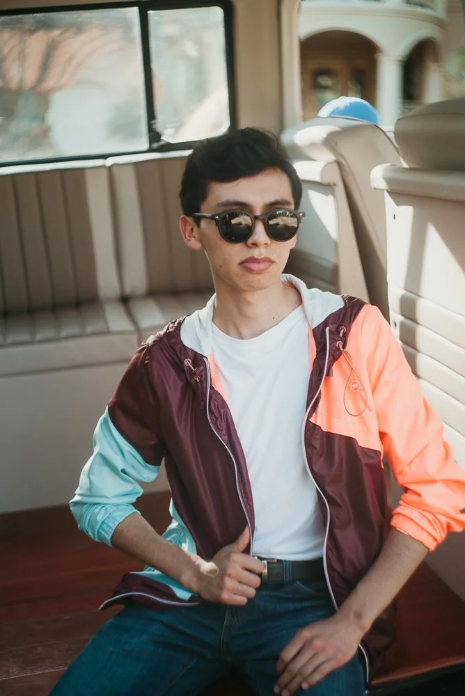 Person with short dark hair wearing sunglasses, a white t-shirt, and a colorful windbreaker posing inside a vehicle.