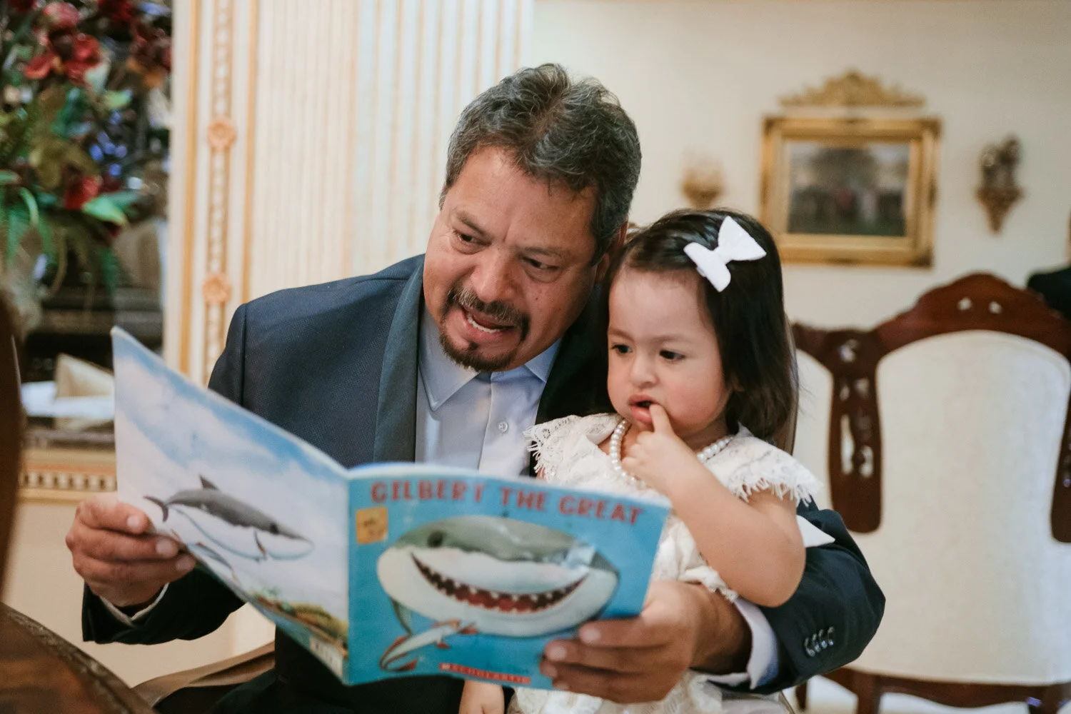 Man in a dark suit reading a children's book titled "Gilbert the Great" with a young girl in a white dress and bow in her hair, sitting together in a decorated room.