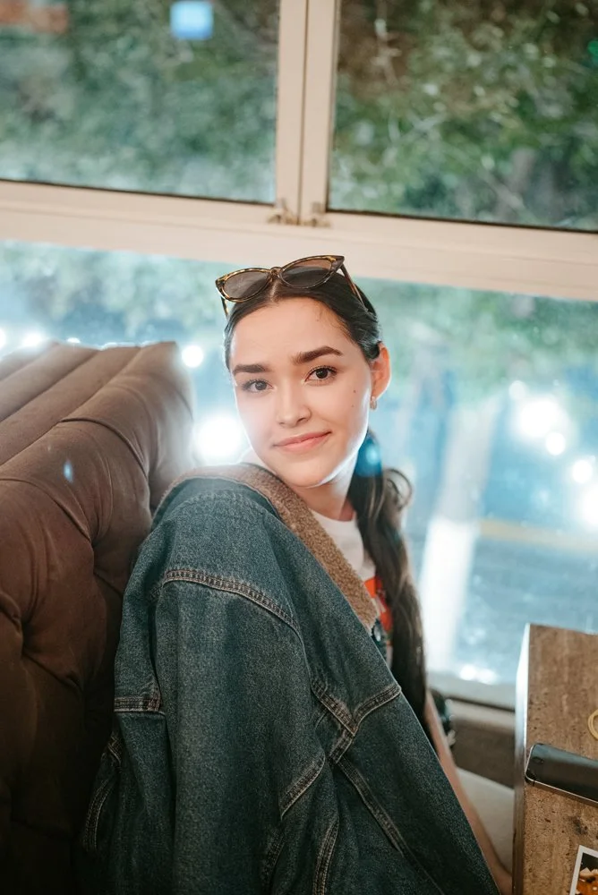 A young woman with dark hair and sunglasses on her head, sitting on a brown cushioned seat in a cafe or restaurant, with a window and trees outside in the background.