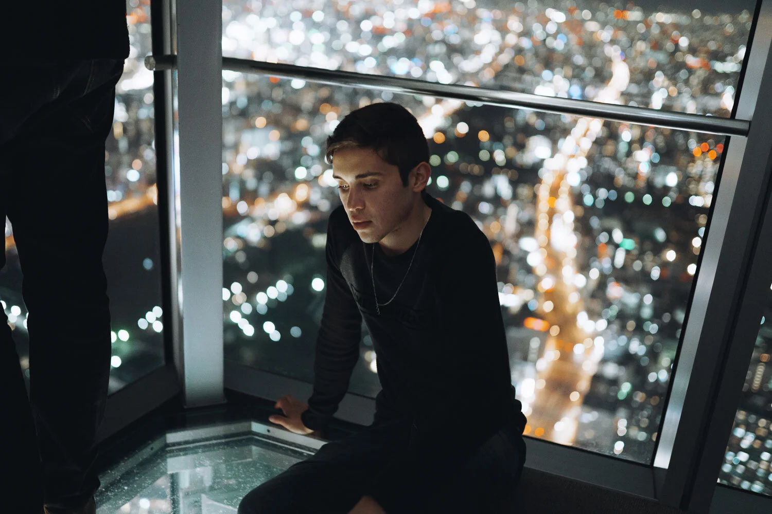 A young man with short dark hair sitting inside a high-rise observation deck at night, looking down with city lights and blurred streets visible through large glass windows behind him.