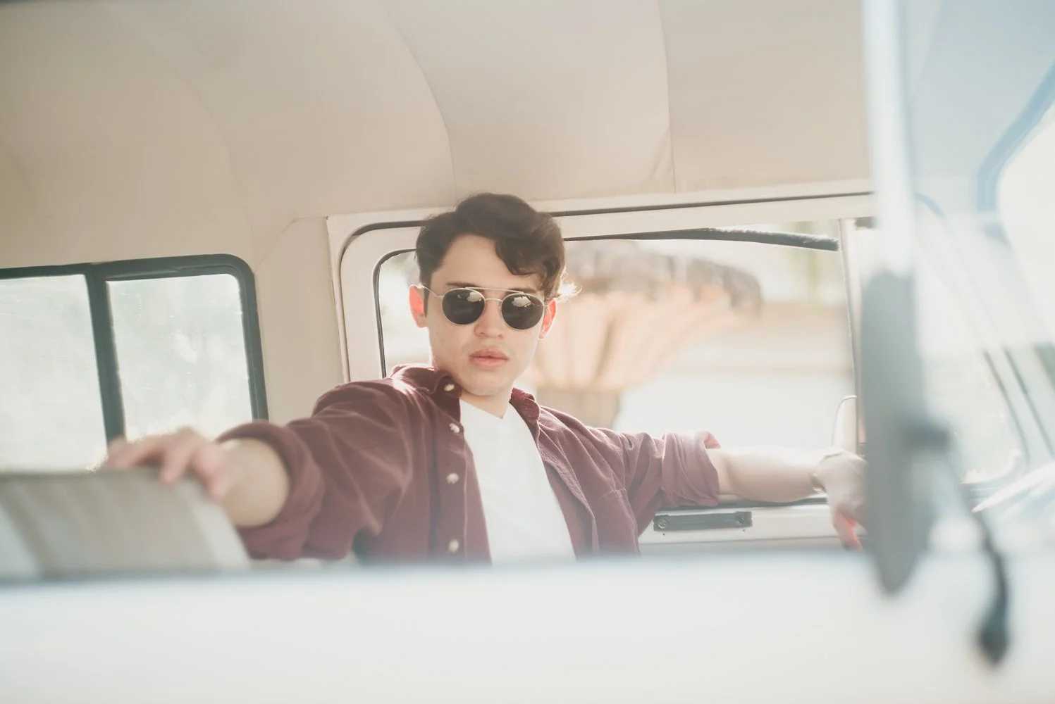 Young man with dark hair wearing sunglasses and a maroon shirt inside a vehicle, extending an arm towards the camera, with a blurred background visible through the window.