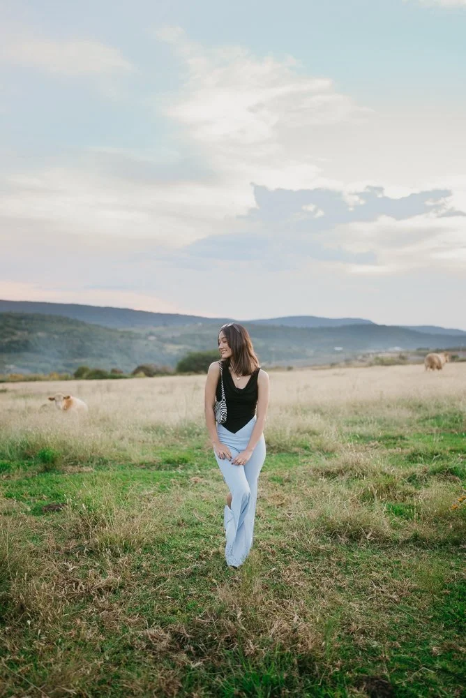 A woman standing in a grassy field with rolling hills in the background under a partly cloudy sky.