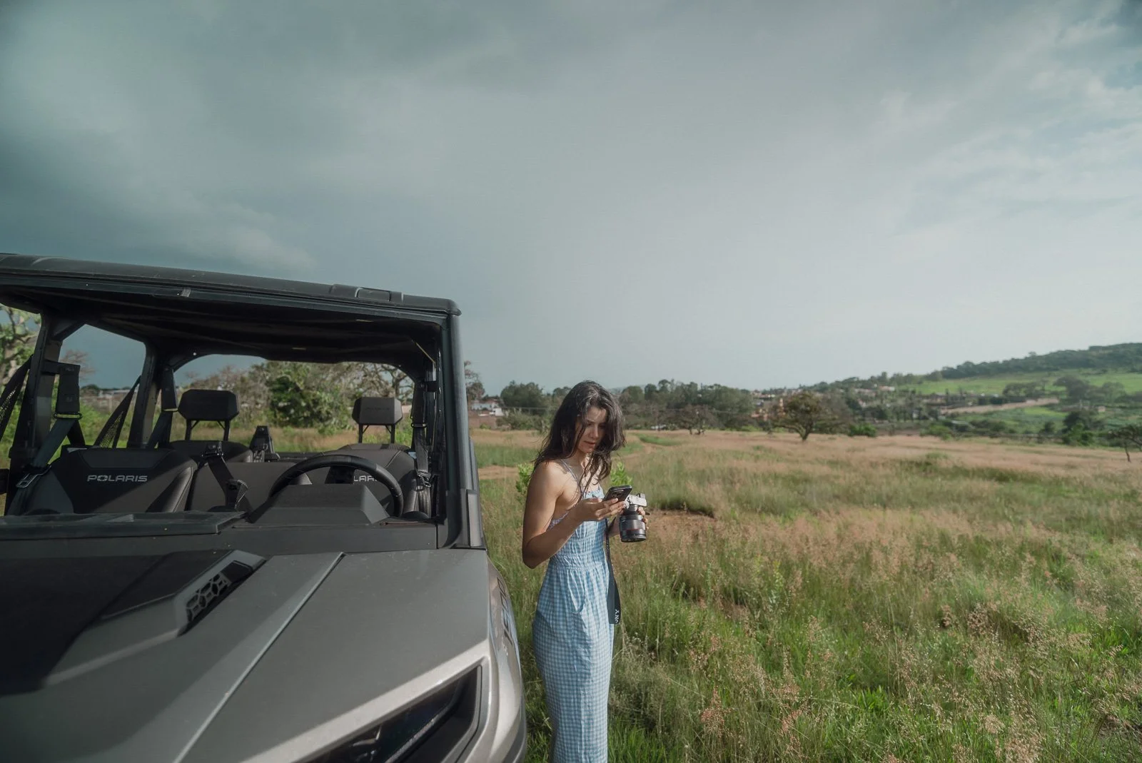 Young woman in a blue checkered dress standing in a grassy field next to an off-road vehicle, looking at her smartphone, with hills and scattered trees in the background under a cloudy sky.
