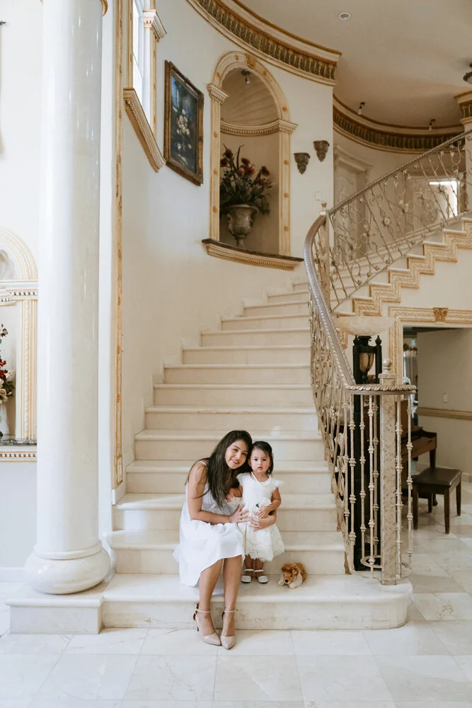 A woman and a young girl sitting on a marble staircase inside a luxurious home, with a small dog sitting beside them.