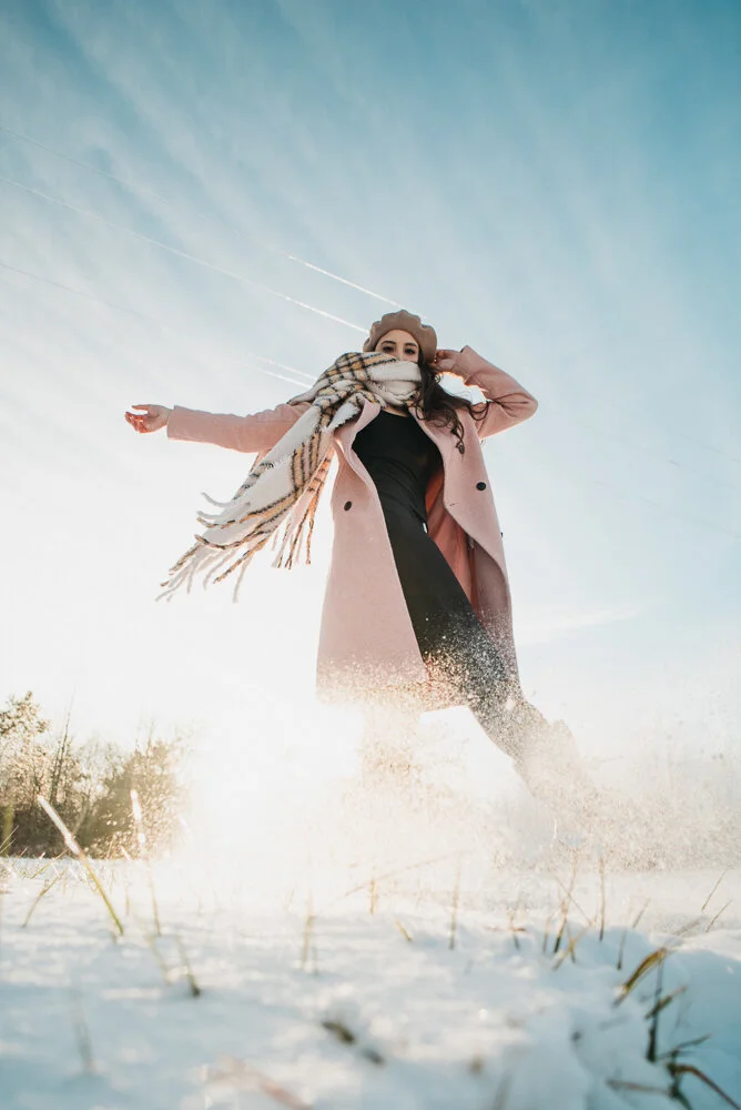 Woman standing in snowy field with arms outstretched, wearing a pink coat, scarf, and hat, with snow kicking up around her, under a clear blue sky.