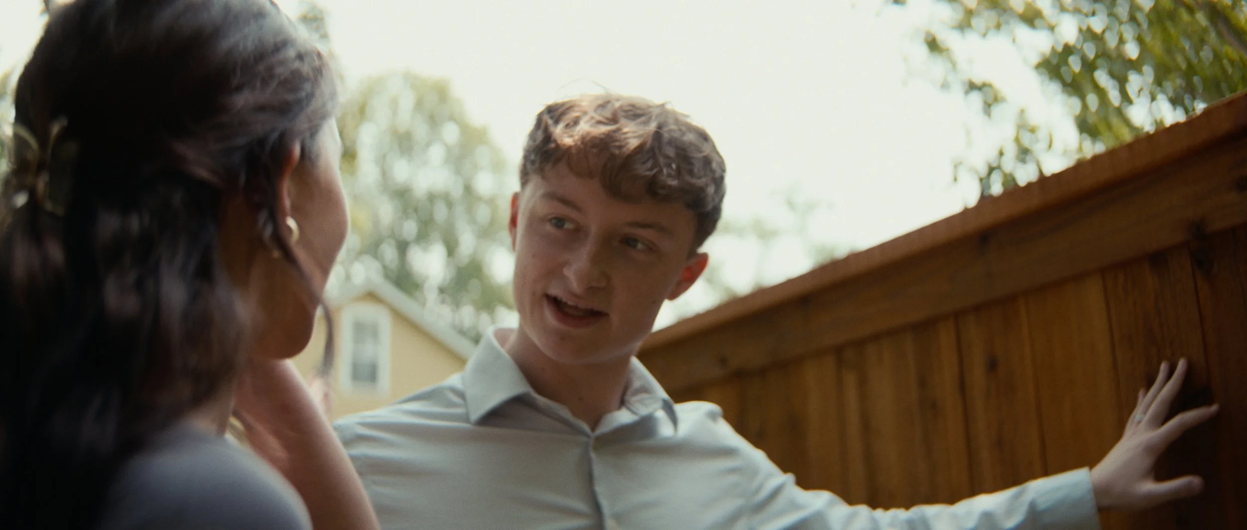 A young man and a woman talking outdoors by a wooden fence on a sunny day.