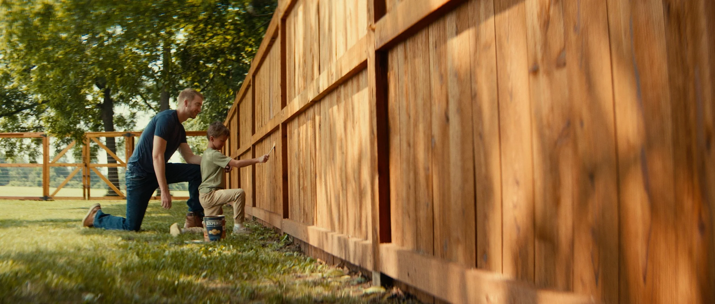 A man and young boy kneeling on grass, painting a wooden fence on a sunny day with trees in the background.