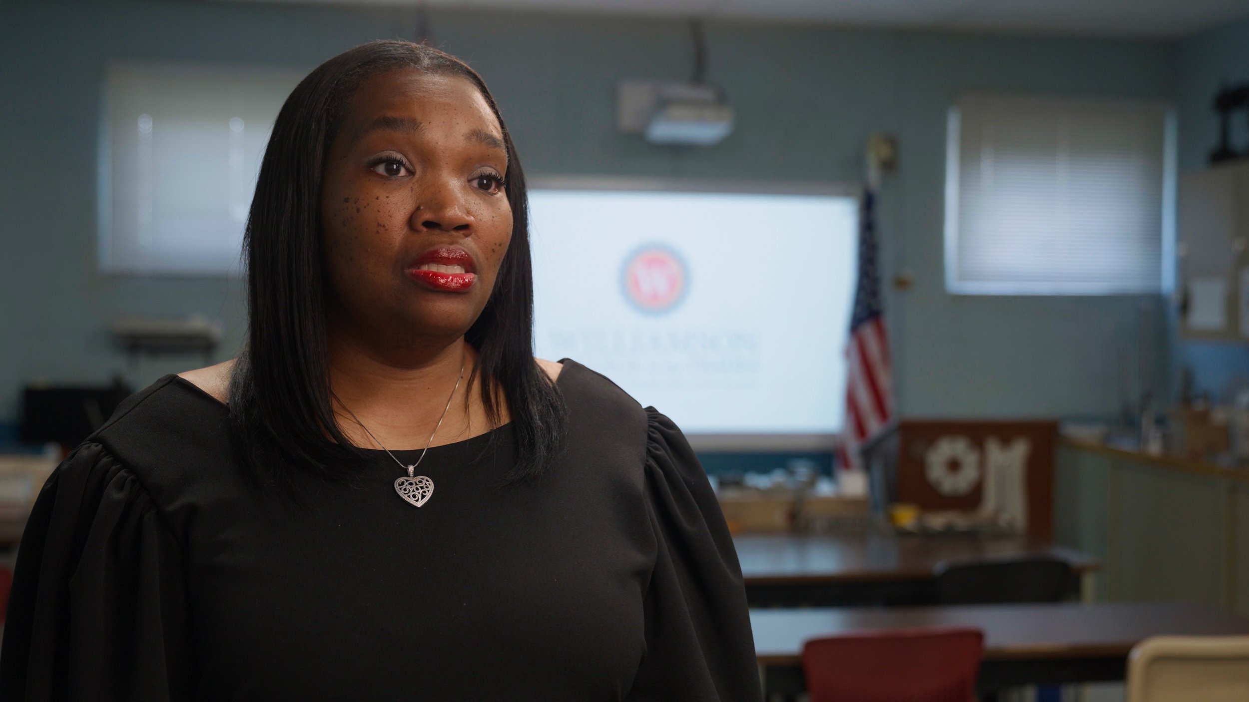 A woman with black hair, red lipstick, and a necklace with a heart pendant, standing in a classroom with an American flag and a projector screen in the background.