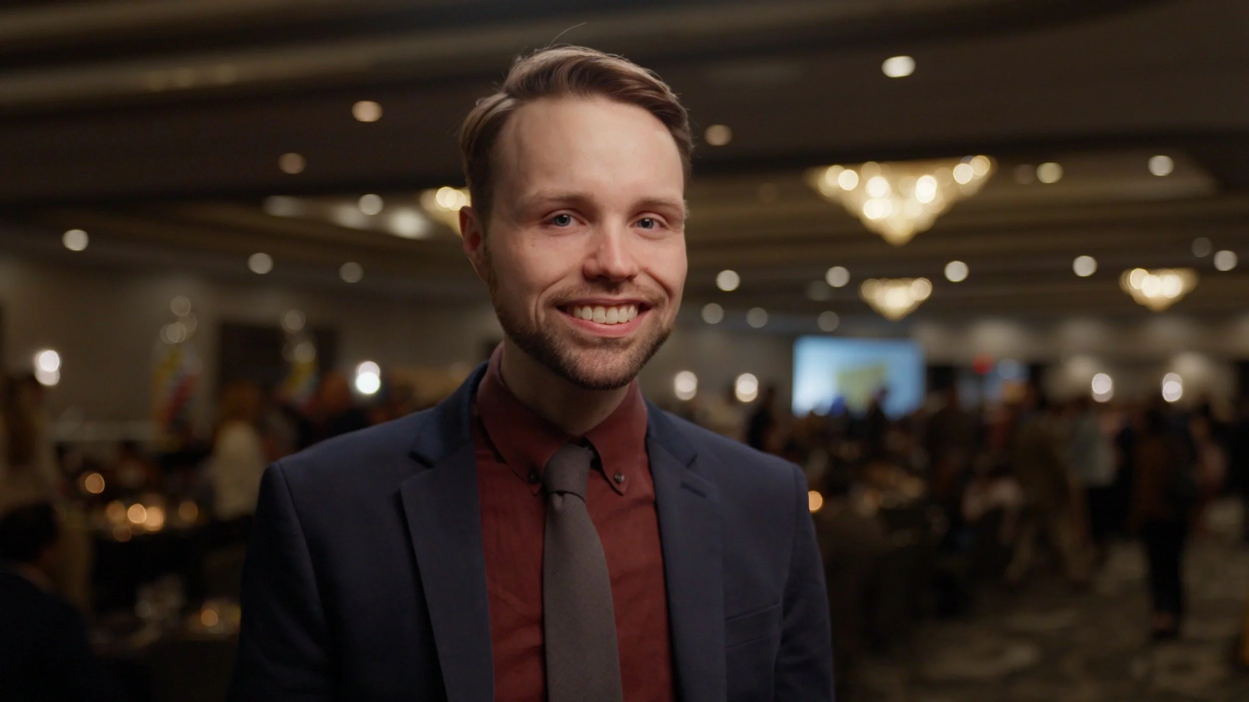 A smiling young man in a suit and tie at a formal event, with a blurred background of chandeliers and other attendees.