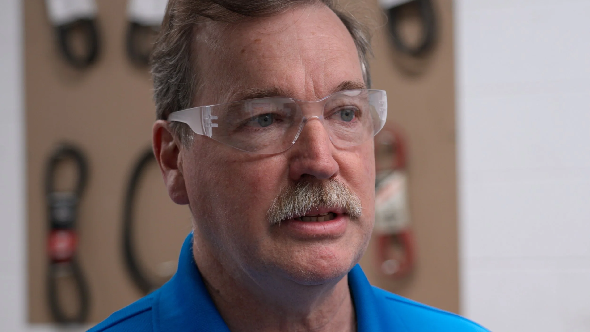 A man with gray hair, mustache, and wearing safety glasses and a blue shirt, speaking in what appears to be a workshop with various tools hanging on a pegboard behind him.