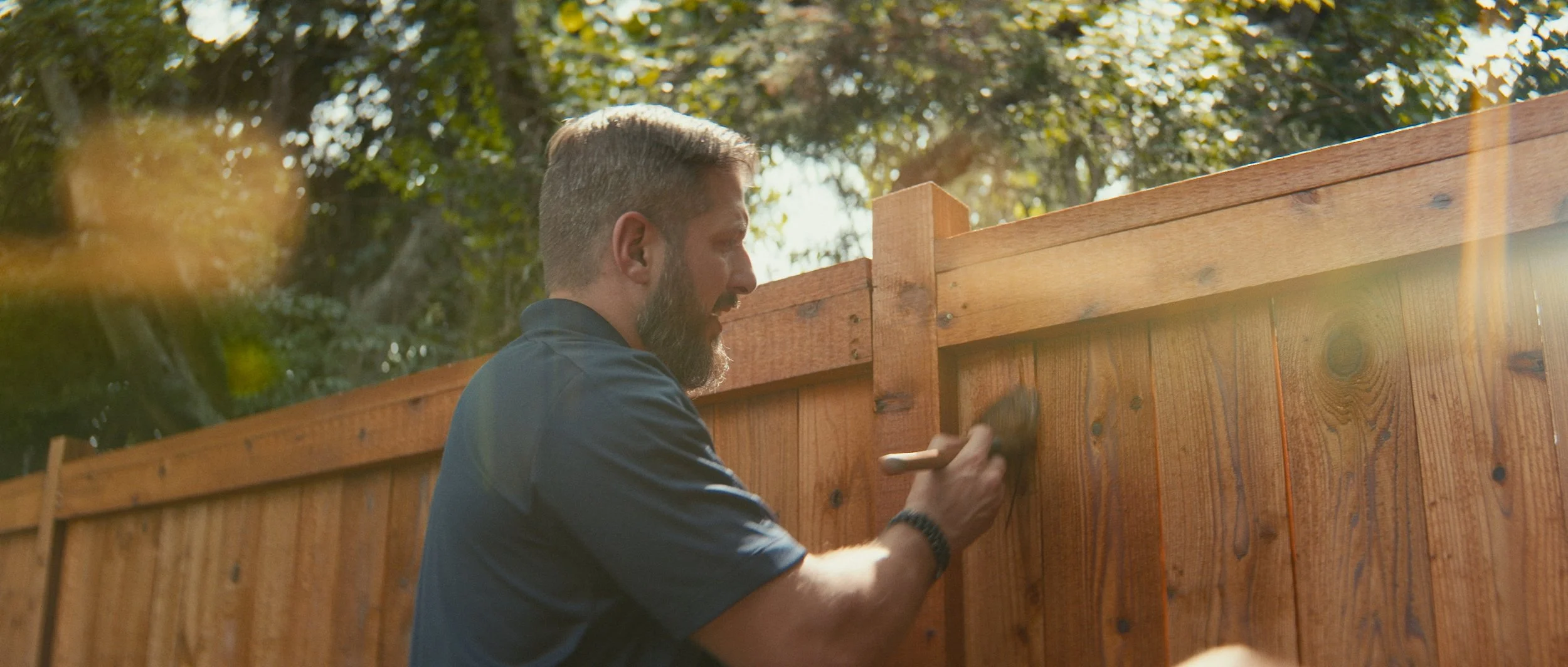 A man with a beard is painting a wooden fence with a paintbrush during daytime.