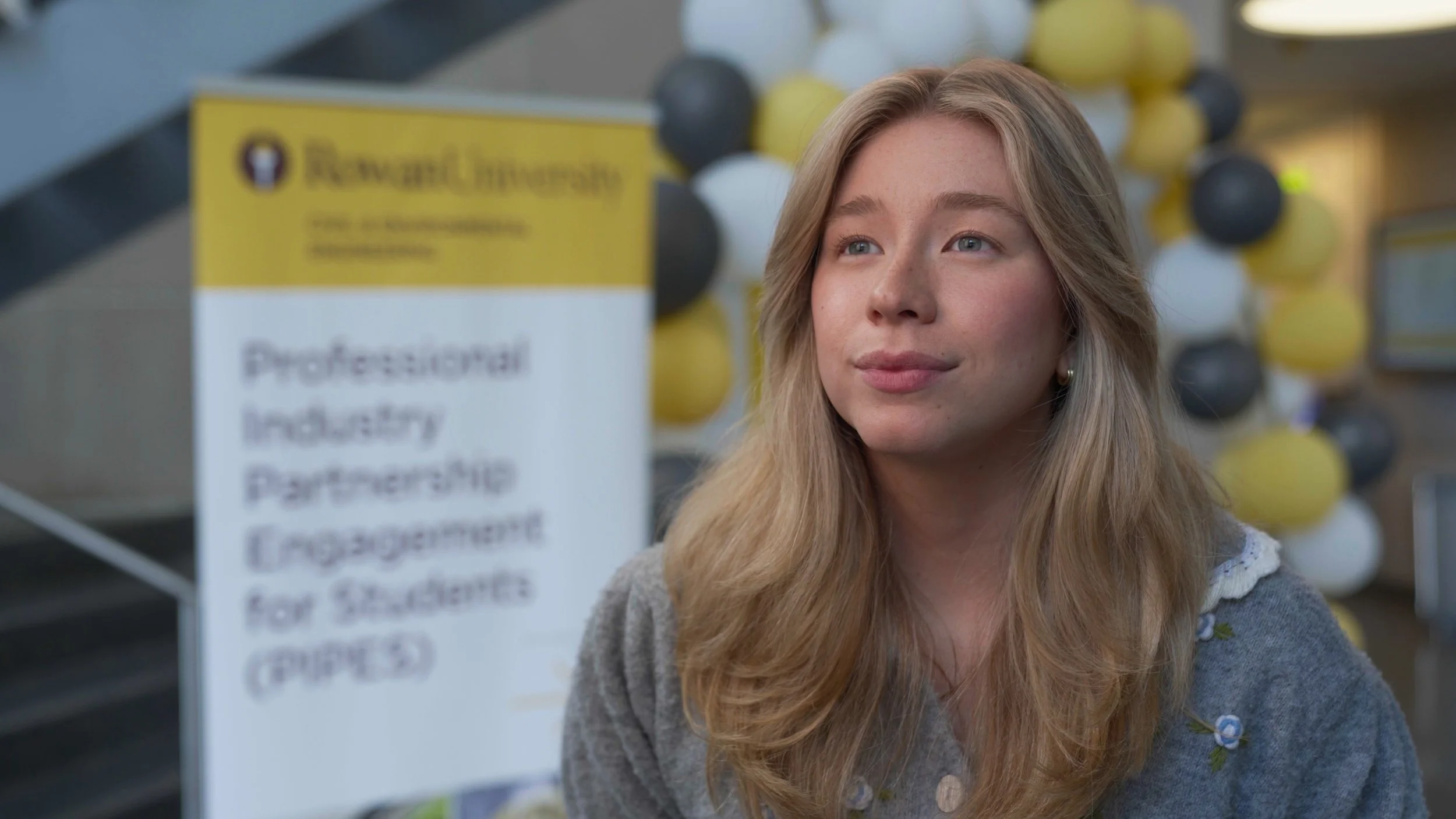A young woman with long blonde hair and a gray cardigan, sitting in front of a yellow and white sign at an indoor event. There are yellow, white, and gray balloons in the background.