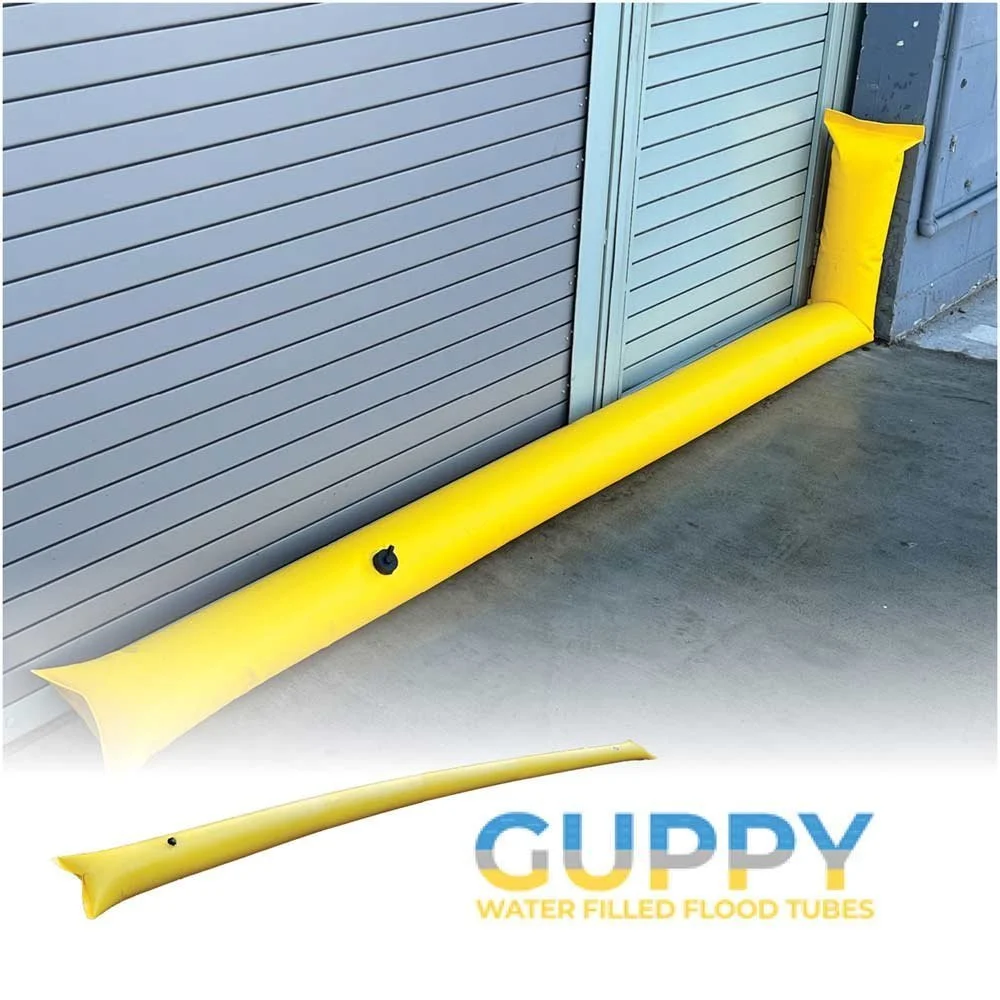 Yellow water-filled flood barrier tube positioned along the base of a metal door and wall. The image also shows a small inset picture of the same tube laid out flat, and a logo with the text "GUPPY WATER FILLED FLOOD TUBES."