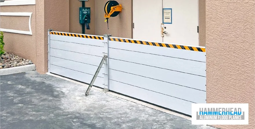 A white aluminum flood barrier gate installed outside a building with beige stucco walls and a gray asphalt driveway.
