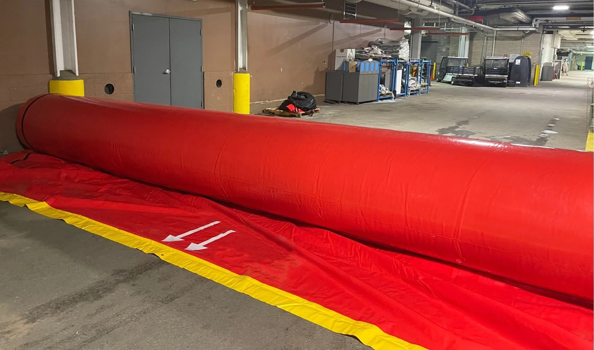 A large red and yellow inflatable mattress or barrier in an indoor parking garage. Various tools, carts, and equipment are visible in the background along with a golf cart and some wrapped items.