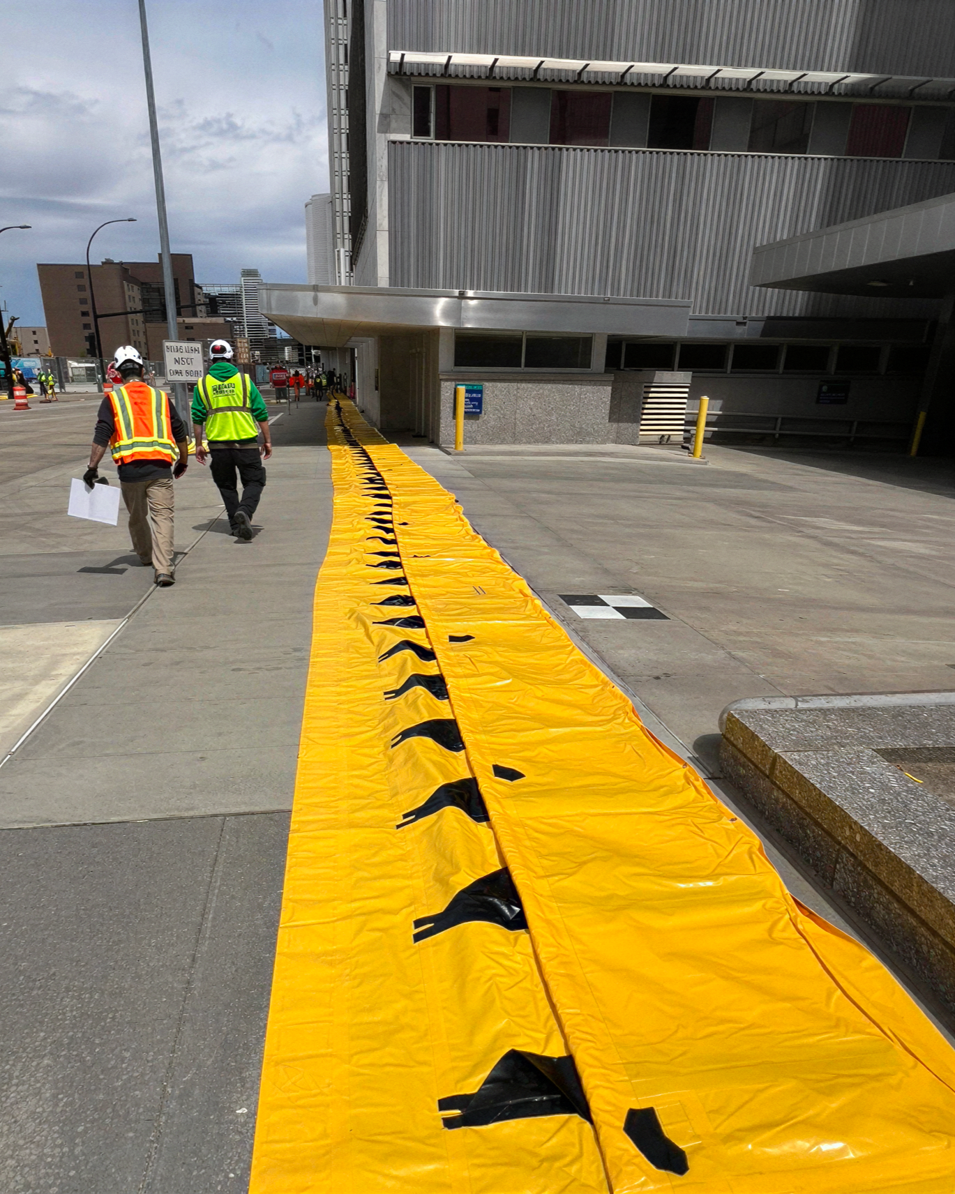 Construction workers walking along a yellow emergency blanket on a city sidewalk outside a building with metallic siding. Some workers are wearing hard hats and safety vests.