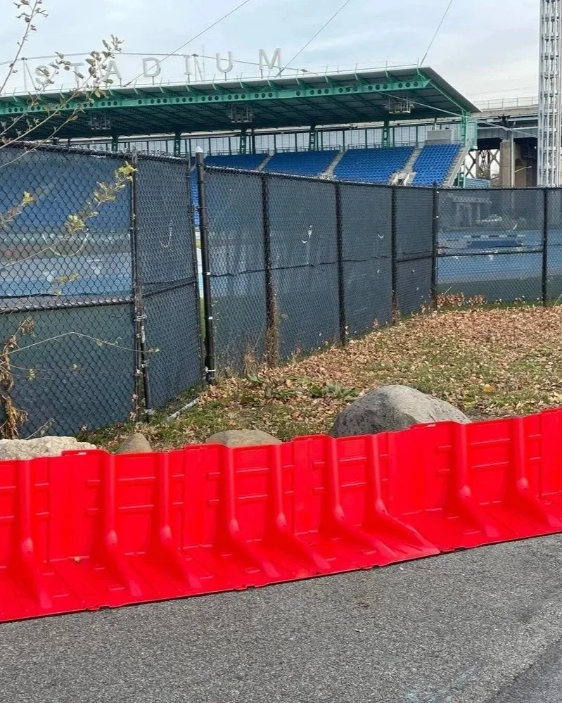 View of a baseball stadium with seating and a fence, partially blocked by a red construction barrier, and some rocks and fallen leaves in the foreground.