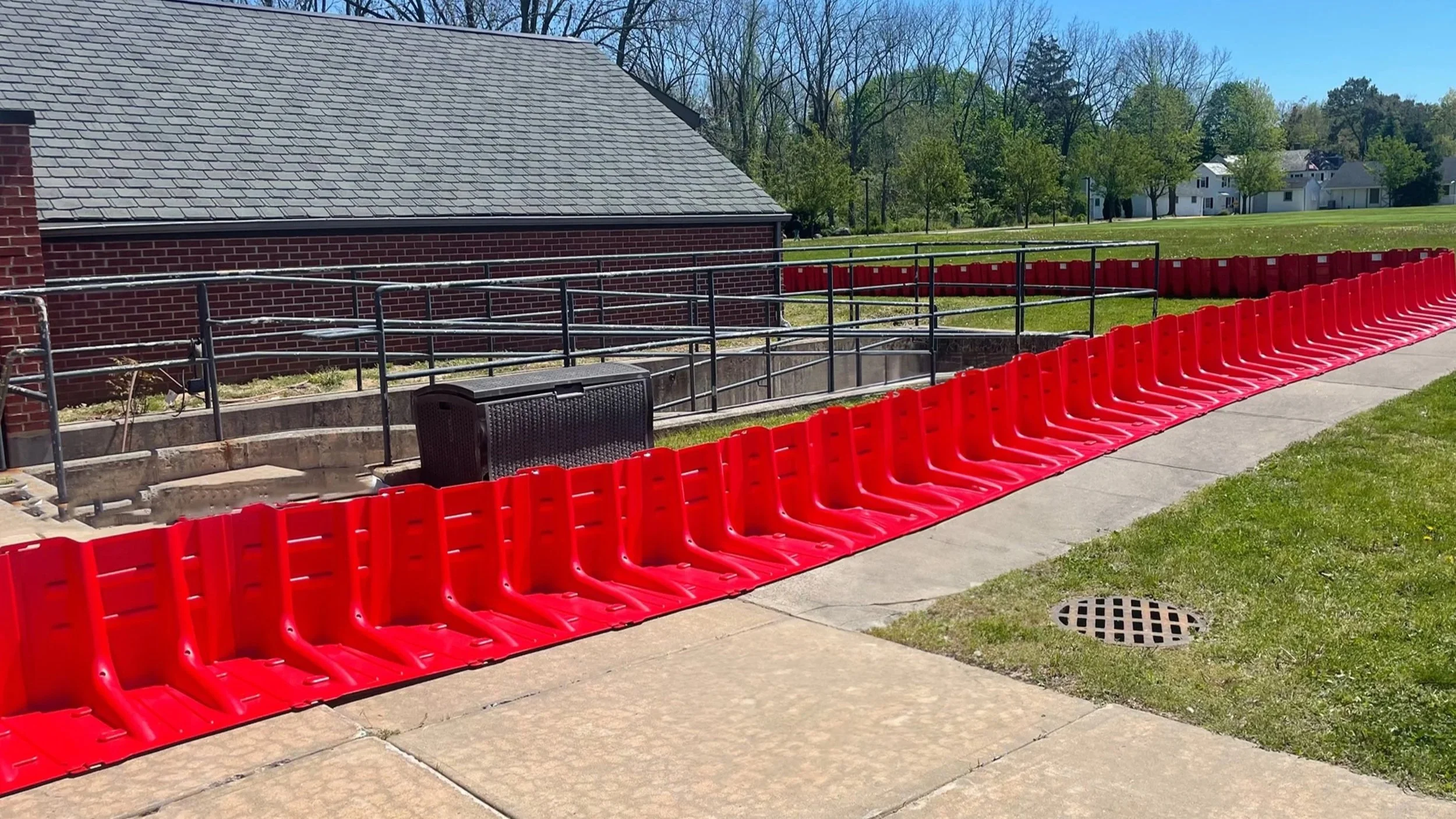 Construction area with red safety barriers, a concrete sidewalk, black railing, a building with a gray shingled roof and brick walls, and a green grassy field with trees and houses in the background.