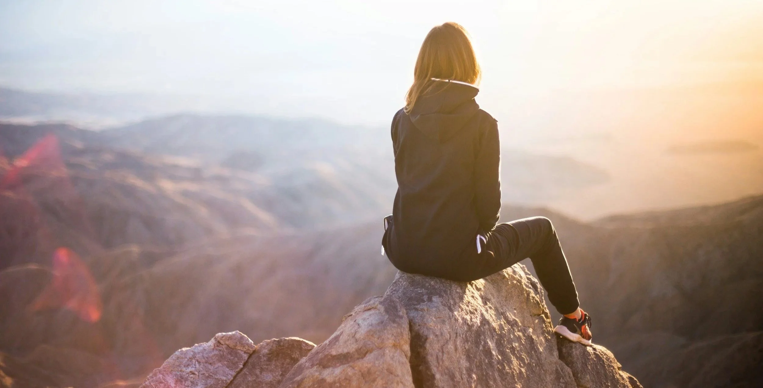 A woman sitting on a rocky outcrop overlooking a mountain landscape at sunset.