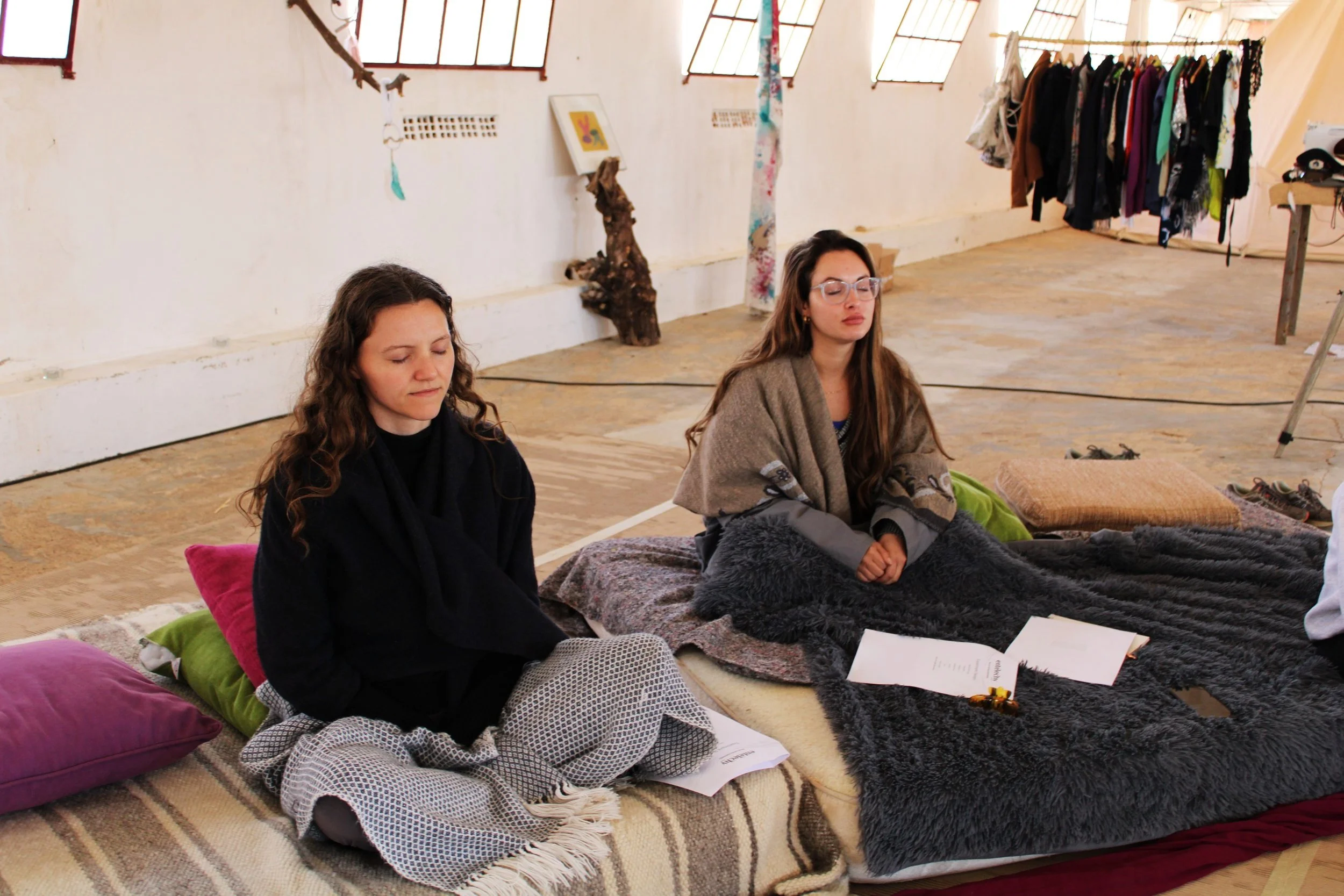 Two women sitting cross-legged on a large carpeted floor, with cozy pillows behind them, in a spacious room with large windows and clothing hanging from a rack in the background. They appear to be meditating or in a moment of calm.