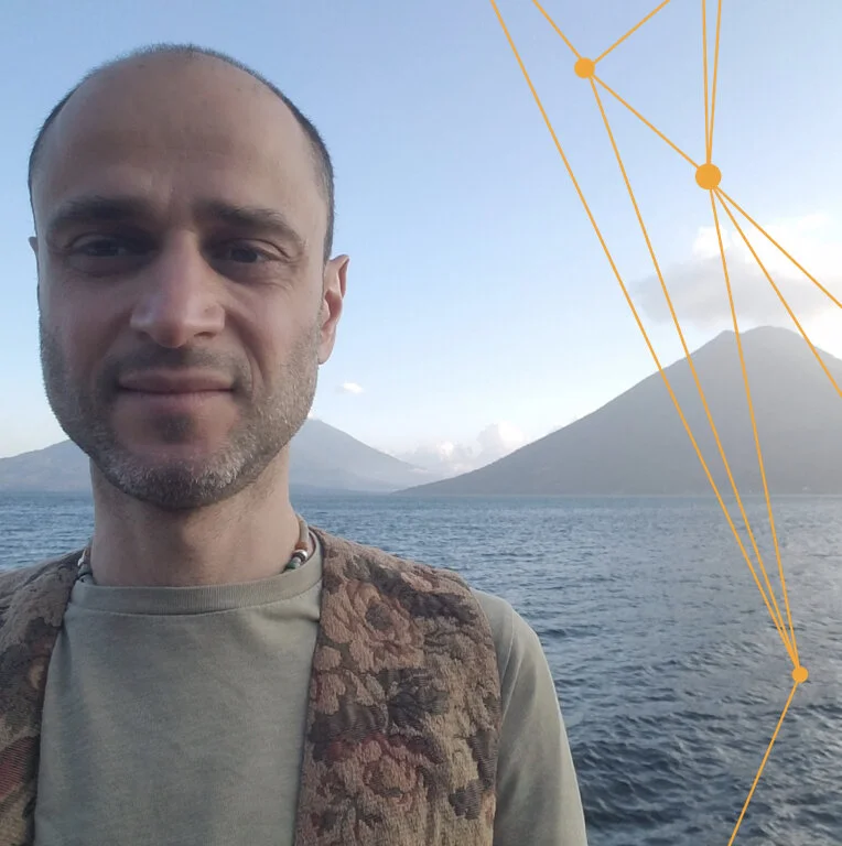 A man with short hair and light facial hair taking a selfie outdoors by a lake with mountains in the background, under a blue sky with some clouds.