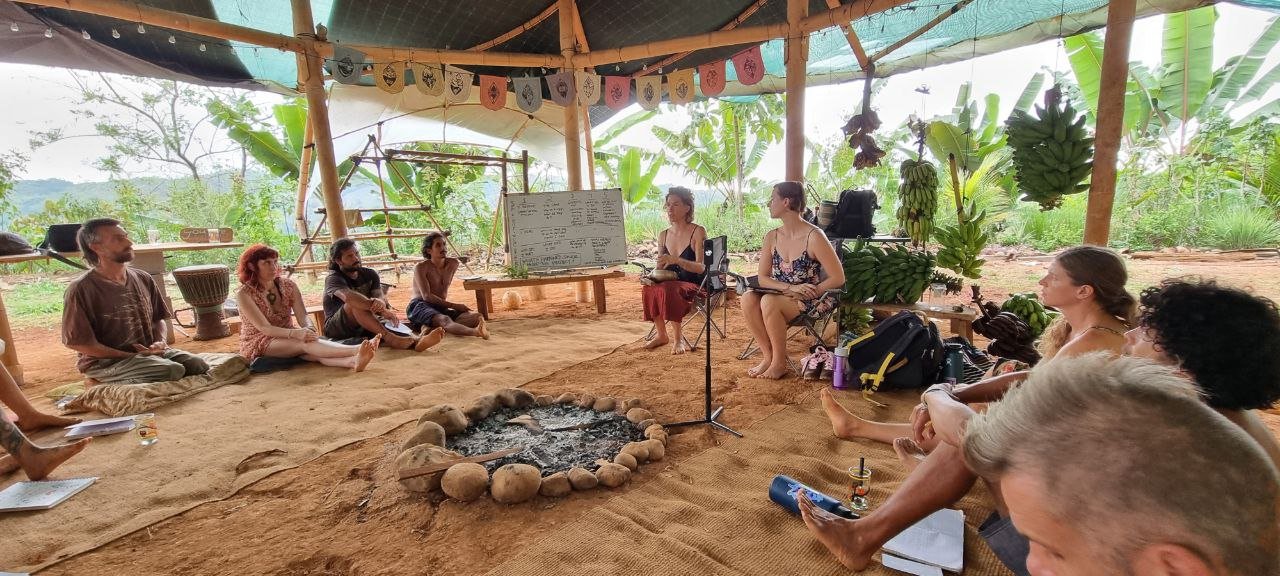 Group of people sitting cross-legged indoors in a circle on natural dirt floor, engaged in discussion or presentation. The space is open with green tropical plants outside, covered with a makeshift roof. A whiteboard with writing and hanging decorations are visible, along with bags and personal items.