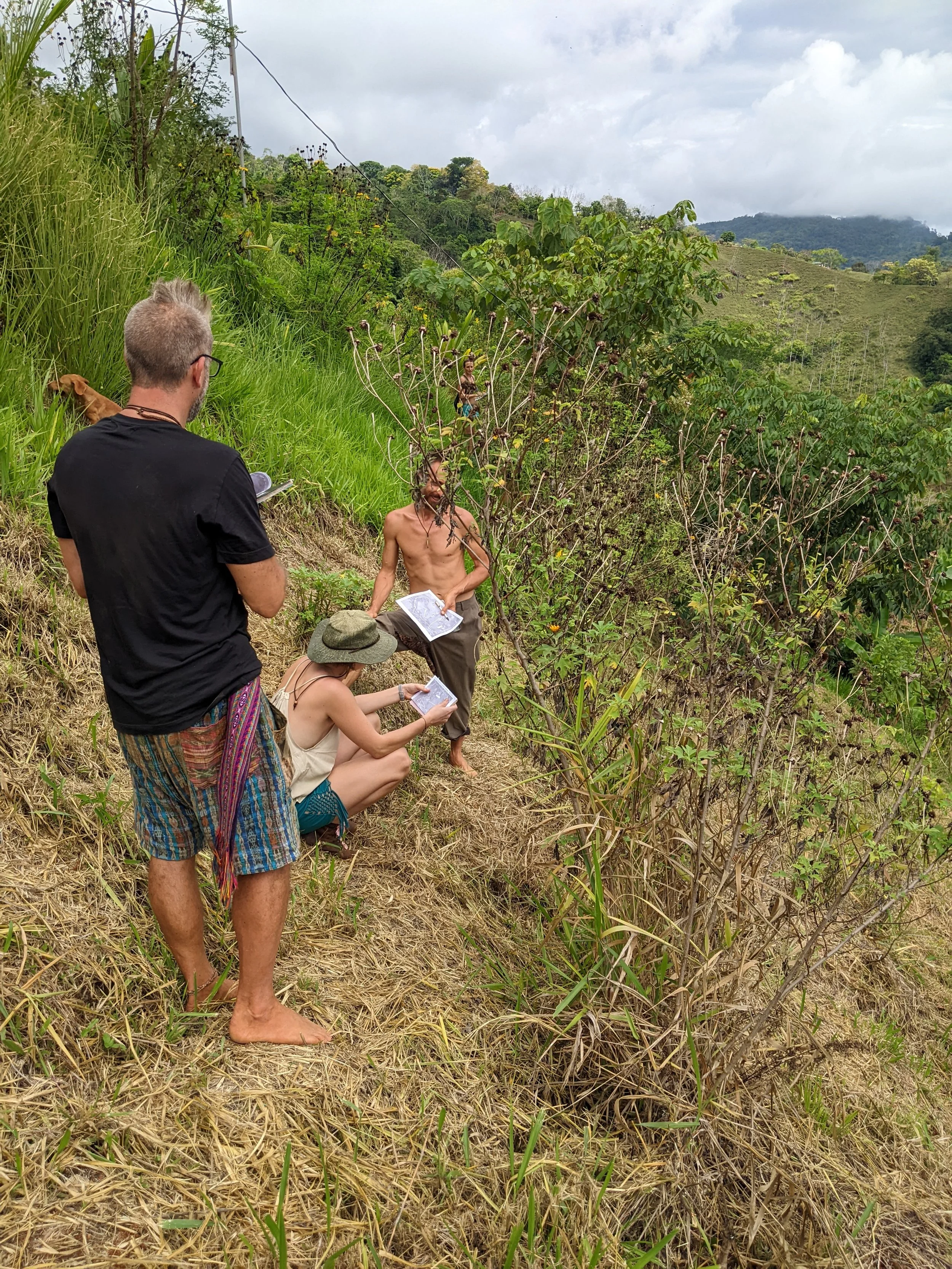 Group of people outdoors in a lush, hilly, and cloudy landscape. One person stands shirtless, holding a paper, with others sitting and standing around him, some with paper or in casual attire, in a natural setting with green plants and dry grass.