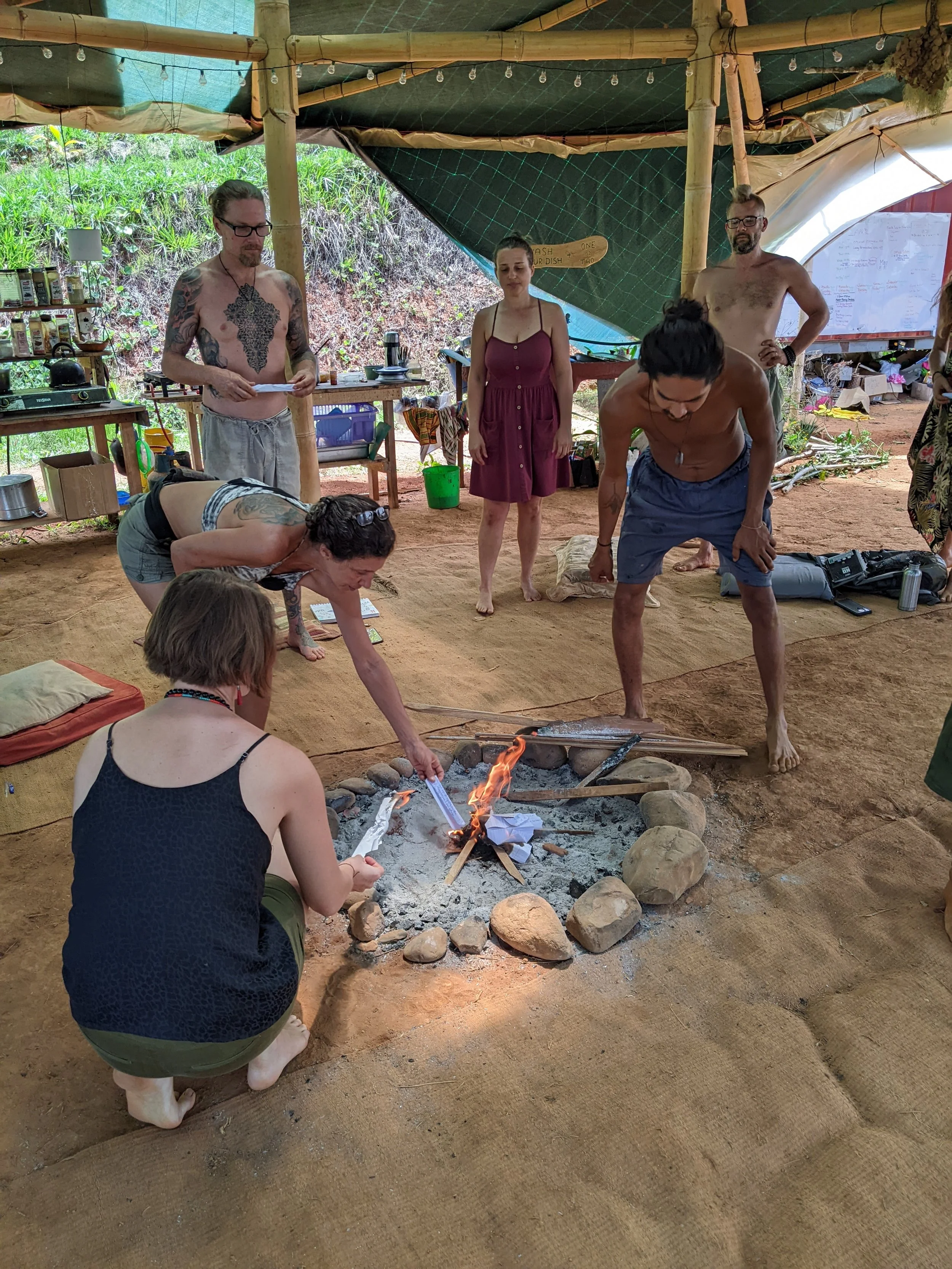 People gathered around a small outdoor fire in a rustic, open-air space with bamboo and fabric roofing. Some individuals are squatting or standing, observing or participating in an activity involving papers or objects on the ground. The scene appears casual and relaxed, with various items and supplies on tables and shelves around the area.