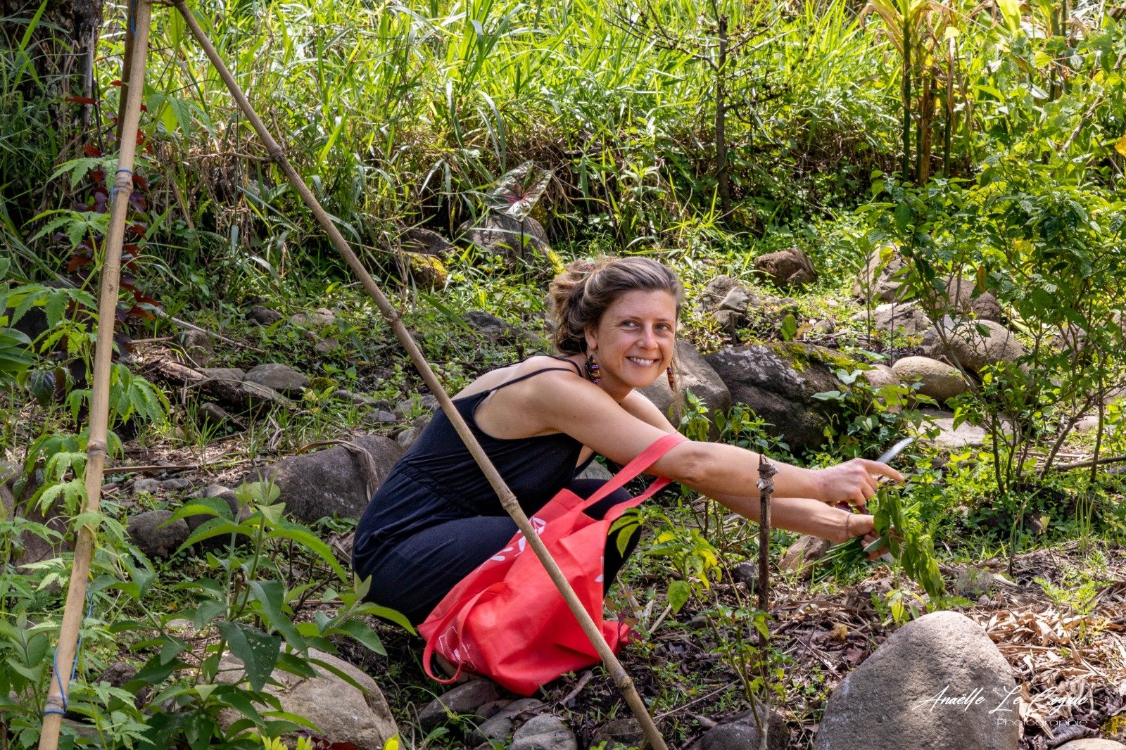 Woman crouching in a lush, green forested area, smiling, wearing a black dress and red bag, surrounded by rocks and dense foliage.