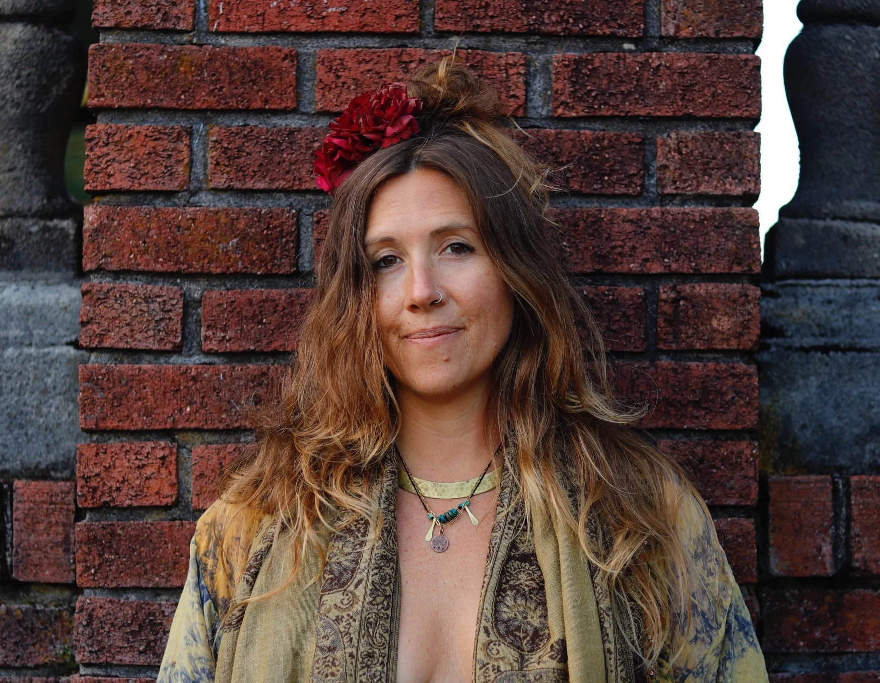 A woman with long, wavy, light brown hair and a nose ring standing in front of a red brick wall, wearing a colorful, patterned jacket and layered necklaces, with a large red flower in her hair.