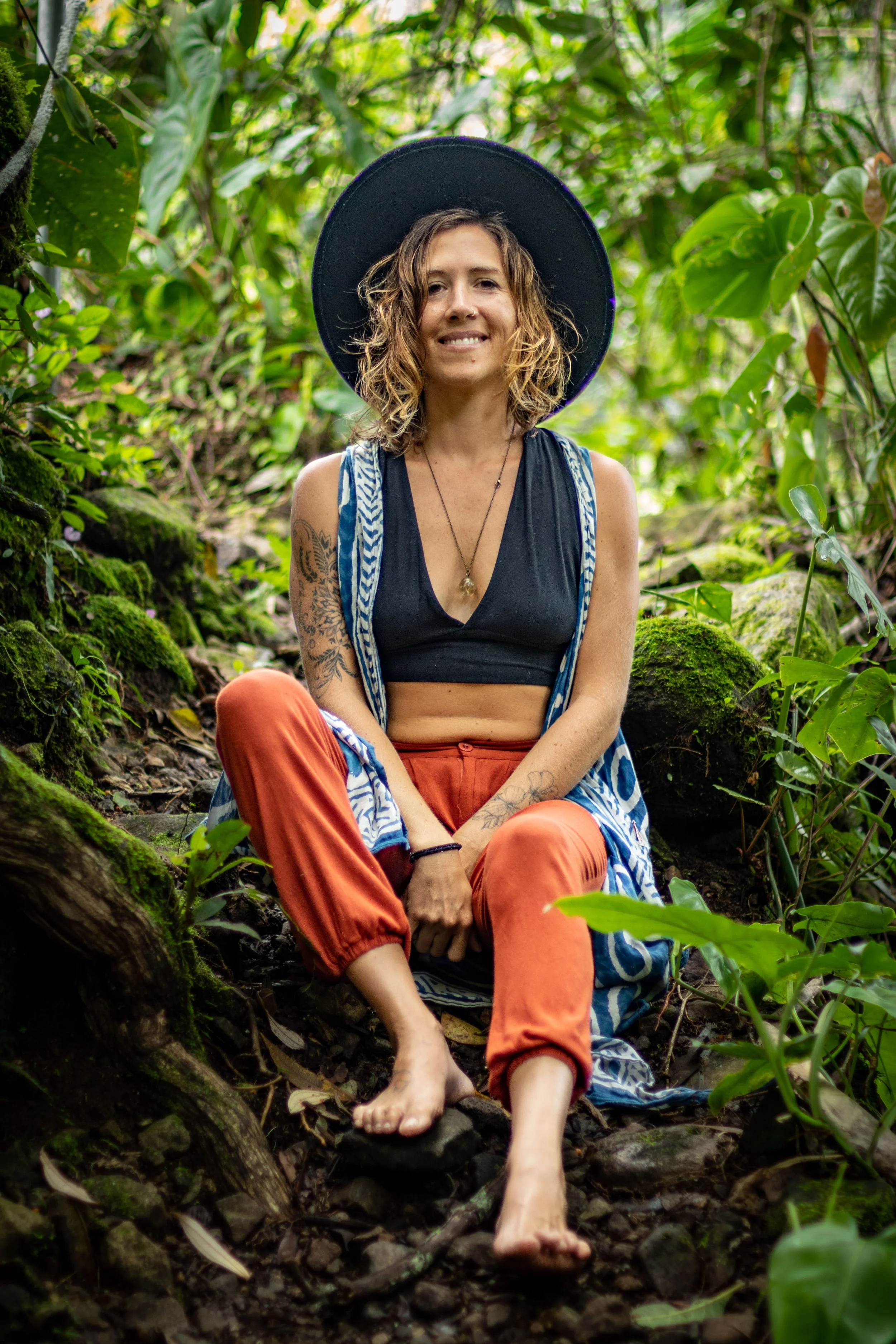 A woman with wavy brown hair, wearing a black hat, black crop top, orange pants, and a blue and white striped robe, sitting barefoot on a mossy log in a lush green forest.