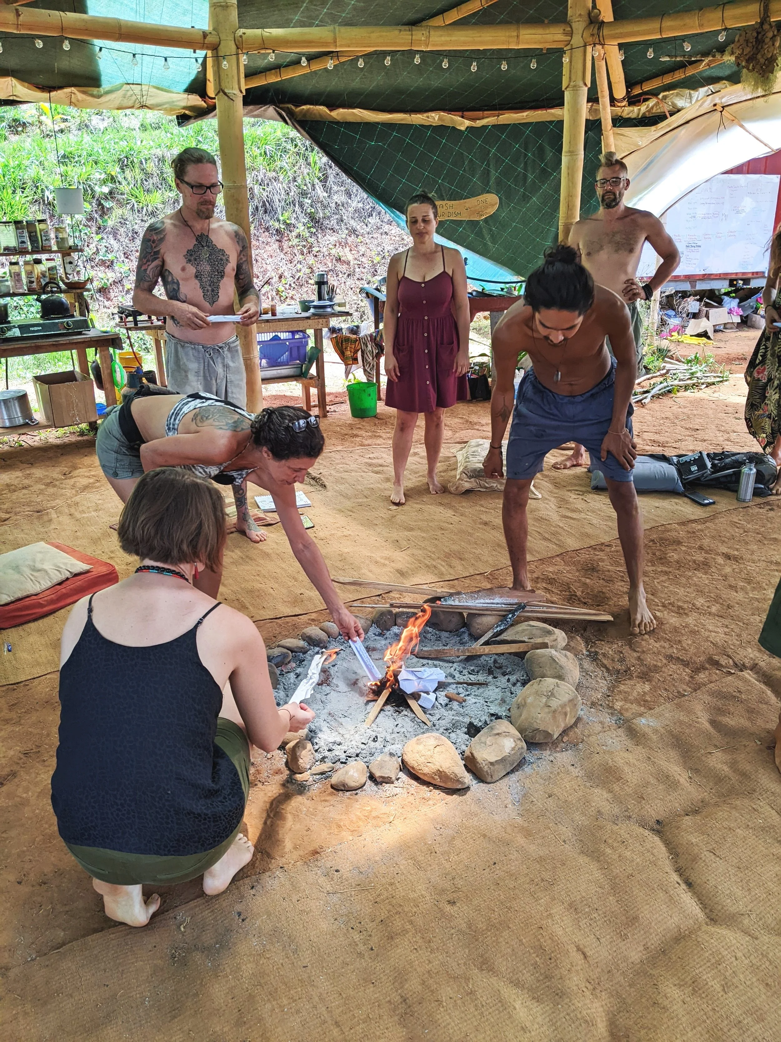 People gathered around a small fire inside a rustic, outdoor shelter made of bamboo and tarps, engaging in a group activity or ritual.
