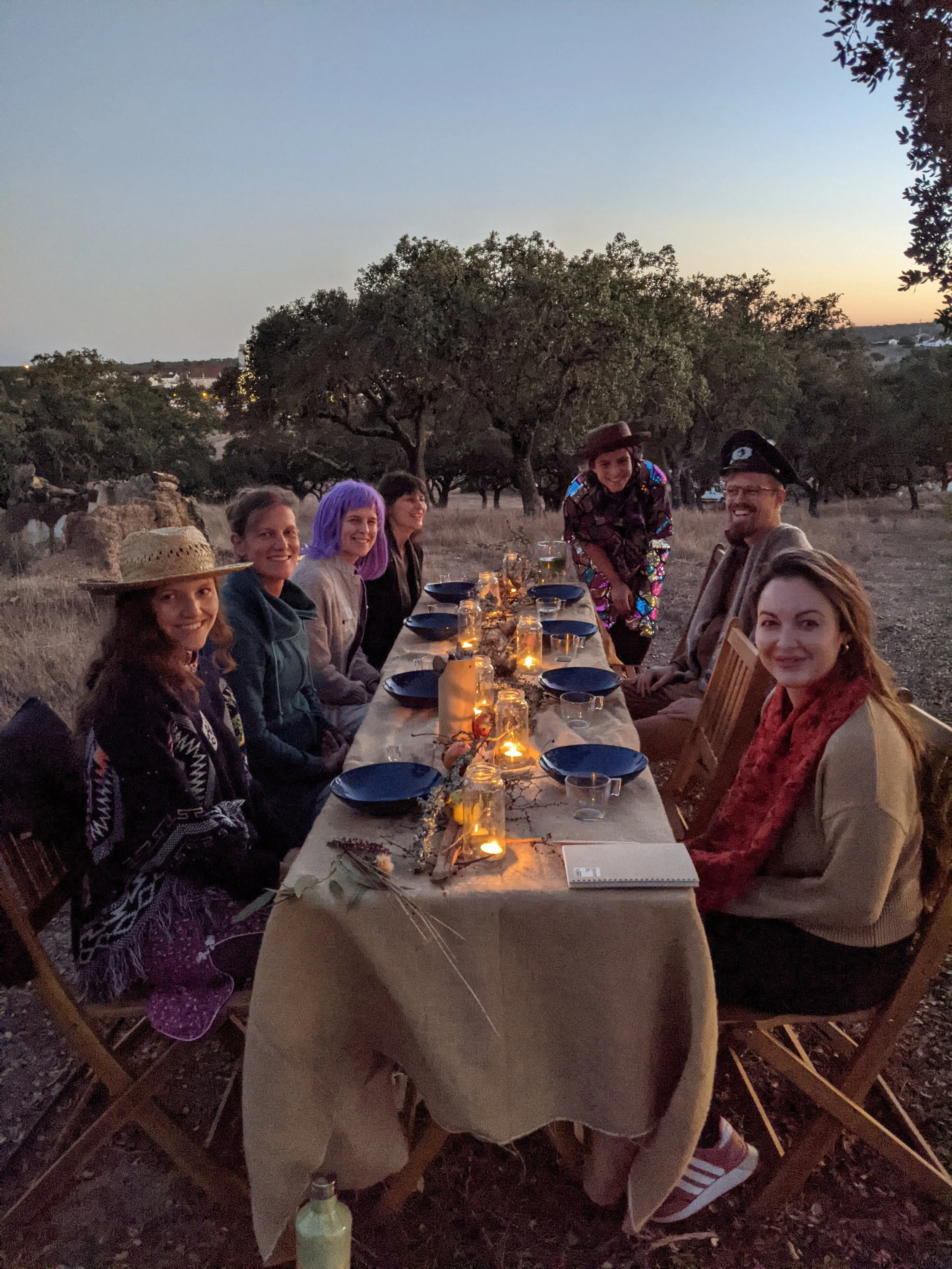 Group of people sitting around a long dinner table outdoors during dusk, decorated with candles and plants, in a natural setting with trees in the background, some with hats and colorful clothing.