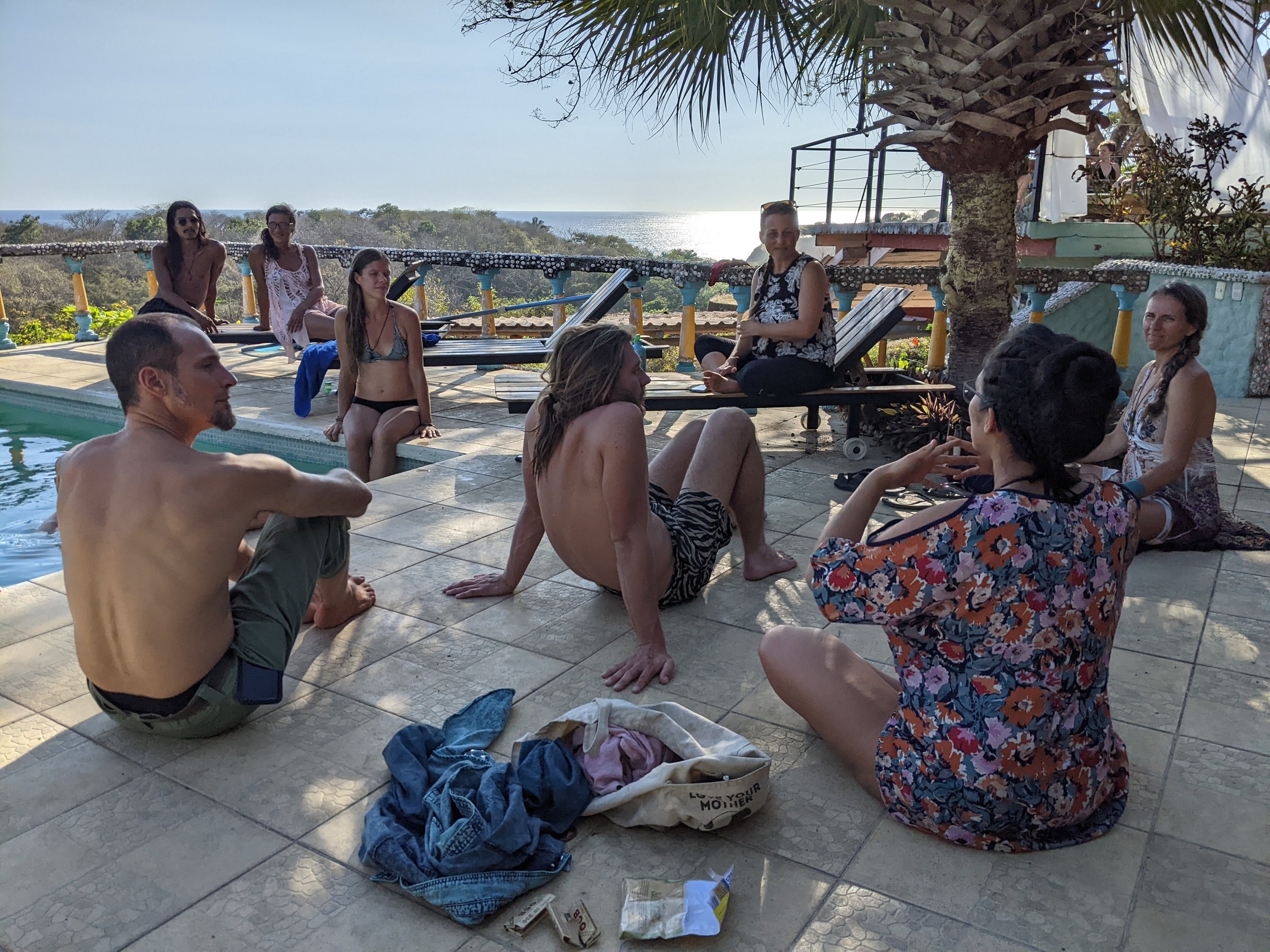People relaxing by a poolside with ocean view in the background, some sitting and some standing, under a tree, during late afternoon or early evening.
