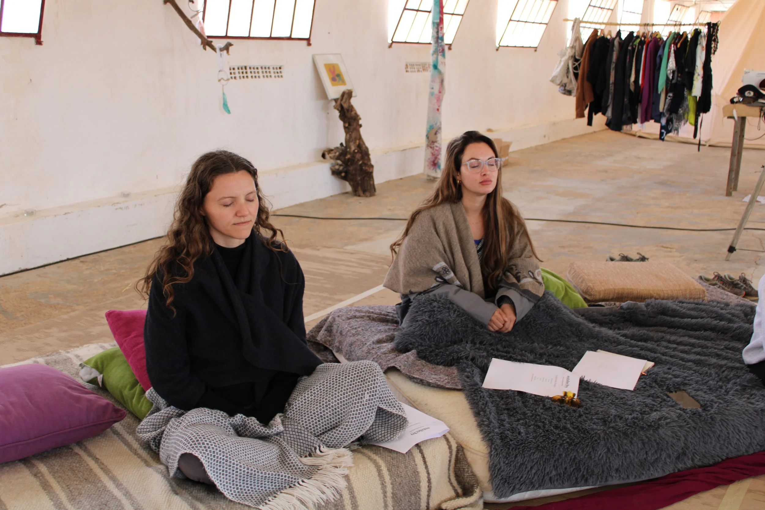 Two women sitting cross-legged on a bed with pillows and blankets in a bright, spacious room with windows and hanging clothing. Both women have their eyes closed and appear to be meditating or practicing mindfulness.
