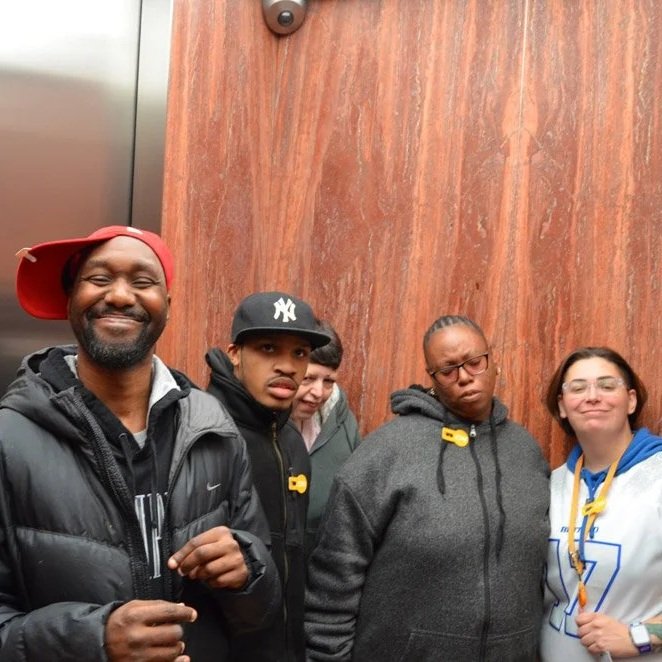 Group of five people standing inside an elevator with a wood panel wall behind them. They are wearing casual clothing, some with identification tags, and appear to be posing for a photo.