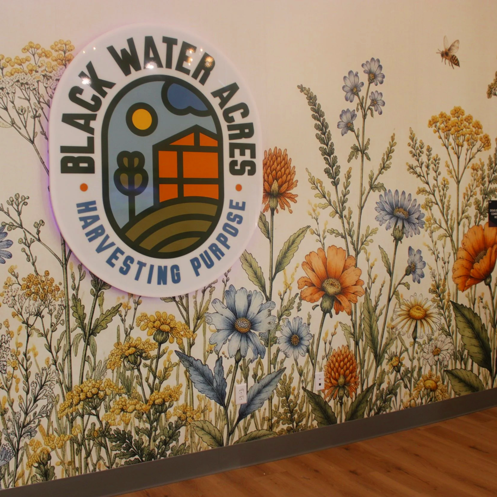 The Black Water Acres logo as a plexiglass sign that is backlit with led lighting on a large full wall floral mural with honeybees on it in the color scheme of oranges, gold, blue, and green.