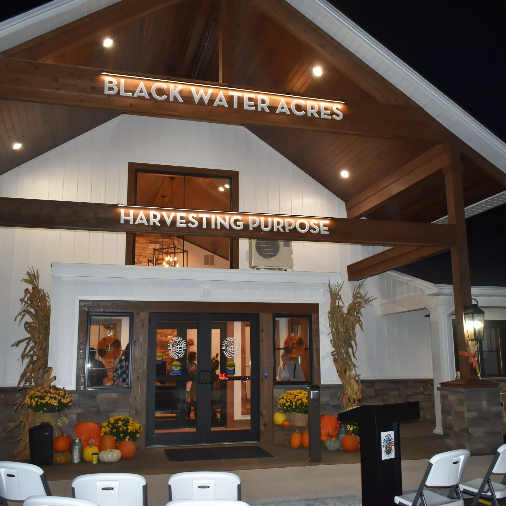 Photo of the front center section of the Black Water Acres building at night with the lights illuminating the building and signage that reads "Black Water Acres" and "Harvesting Purpose".