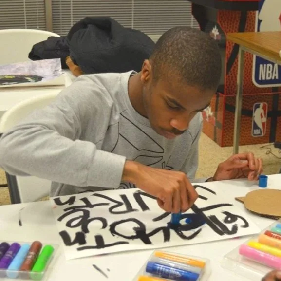 A young man is creating art on a piece of paper with colorful markers, sitting at a table with other children wearing casual clothes.