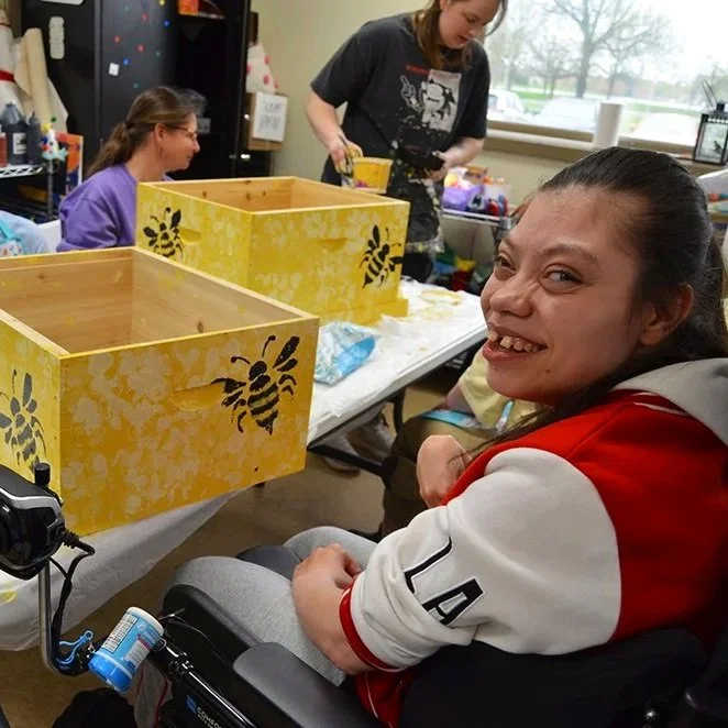 Photo of a young woman in a wheelchair working on yellow bee boxes in our art program. The young woman is wearing a white and red shirt smiling for the photo. There are two additional females in the background of the photo, a staff member, and anothe