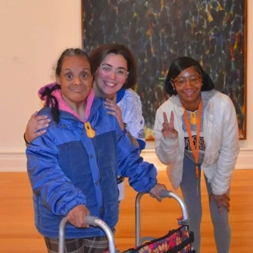Three women smiling at an art gallery, with colorful abstract paintings in the background, one of them using a walker.