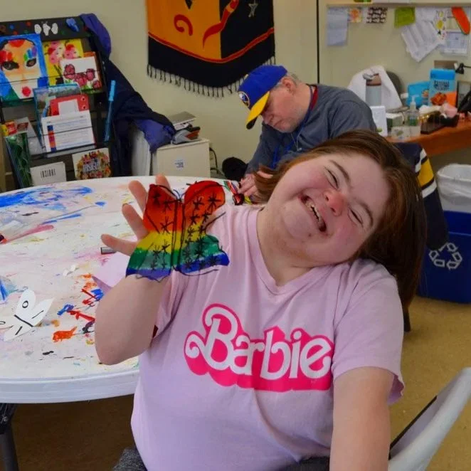 A smiling girl wearing a pink 'Barbie' T-shirt, showing her rainbow-colored hand with a butterfly-shaped glove, sitting at a table covered with colorful art supplies and crafts. A man in the background is working at a table in a room with various ite
