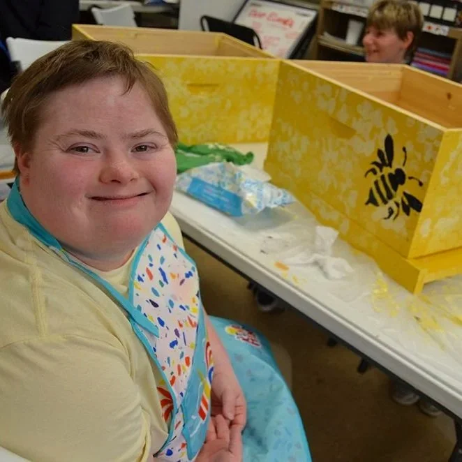 Smiling girl painting bee boxes in our art program for Black Water Acres. She is sitting at a table wearing a paint smock and is painting the bee box yellow with a black bee image on it.