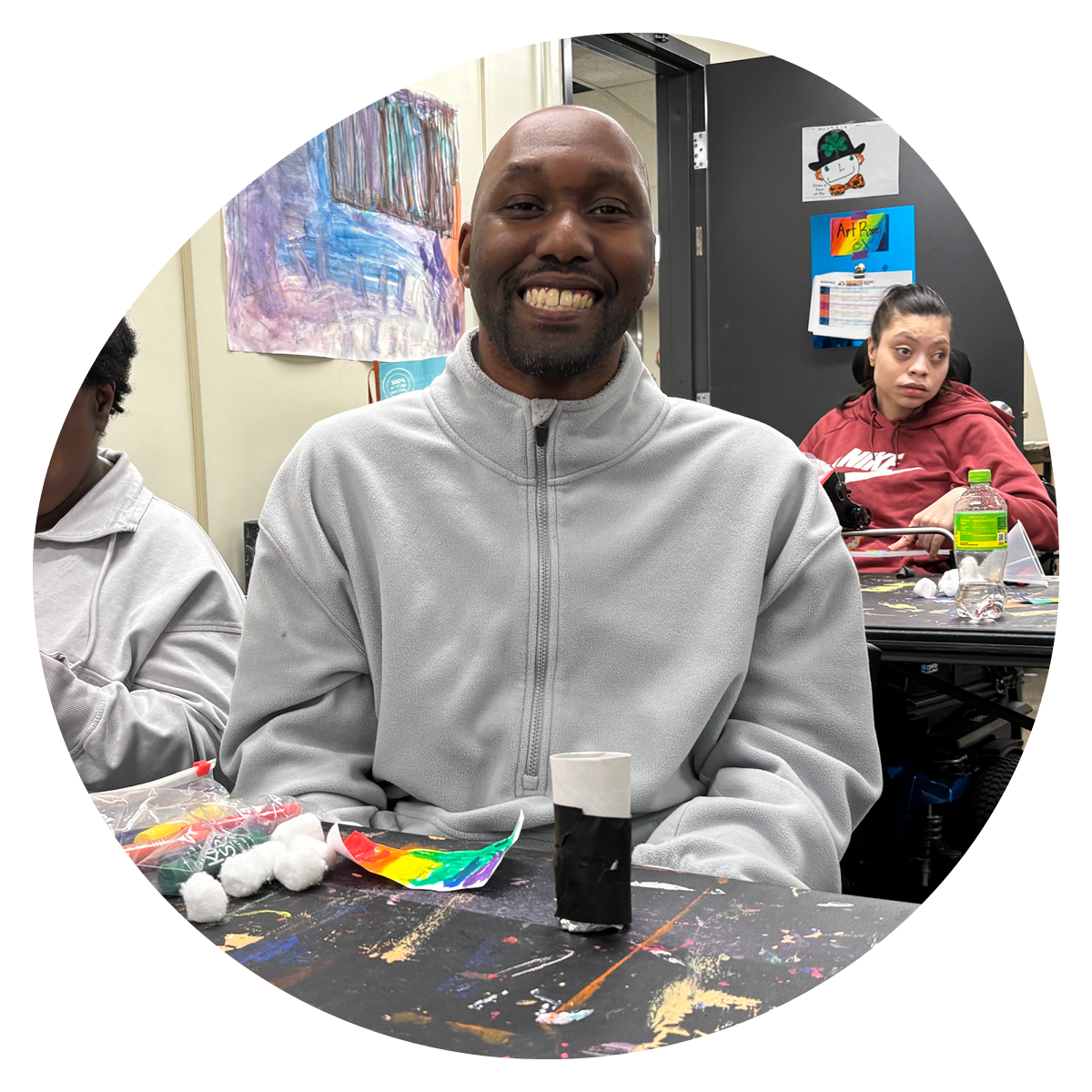 A smiling man sitting at a table with arts and crafts supplies in a classroom, with a young woman in a red hoodie in the background.