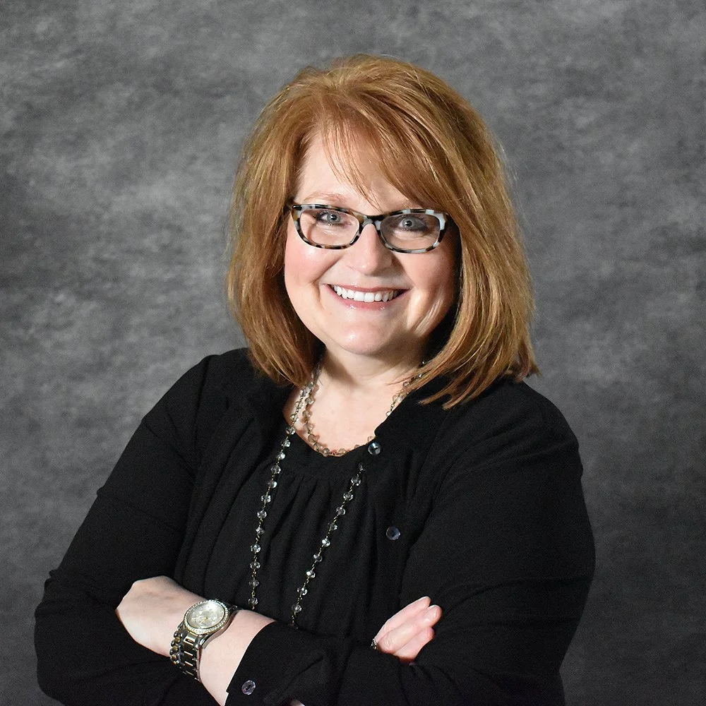 A smiling woman with red hair, wearing glasses, a black top, and a long necklace, standing against a gray background.