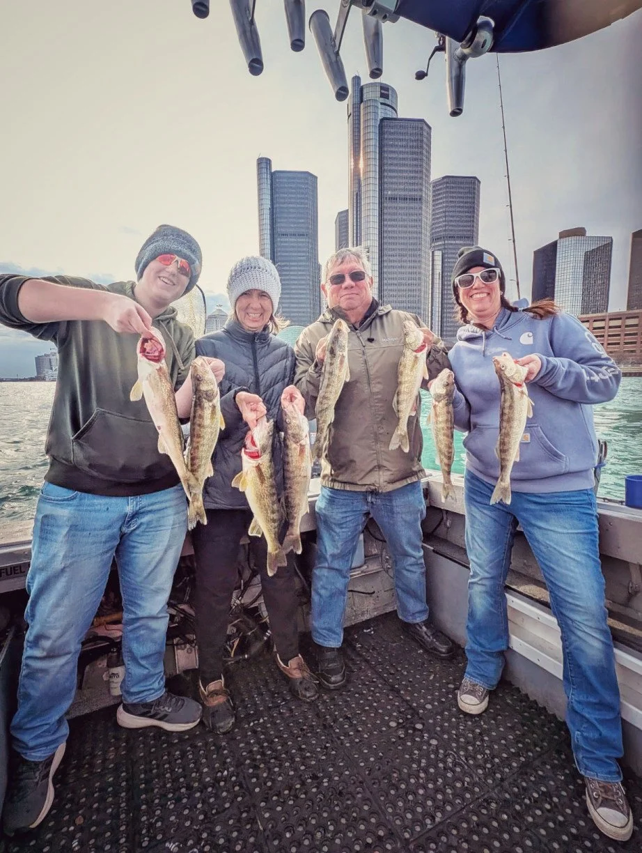  A group of anglers holding up several walleye on a Reel Fish'n charter boat with the Detroit skyline and Renaissance Center in the background. 